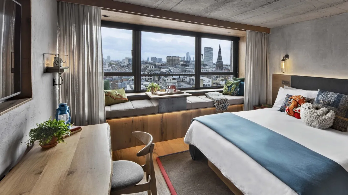 A Treehouse Hotel London room with a cushioned window seat running along a wide window over the West End rooftops and a church spire, a concrete ceiling, a live-edge wooden desk and a bed with a blue throw and a stuffed sloth