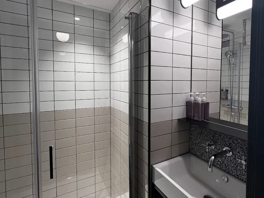 A bathroom at TRIBE London Canary Wharf with a walk-in shower behind a glass door, white and taupe tiles with black grout, and a grey basin under a lit mirror