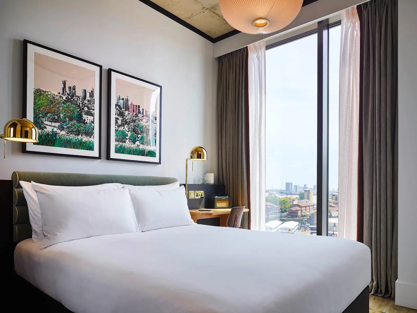 A room at TRIBE London Canary Wharf with a double bed against a green upholstered headboard, brass wall lamps, two framed prints of the Canary Wharf skyline, and a floor-to-ceiling window looking out over east London