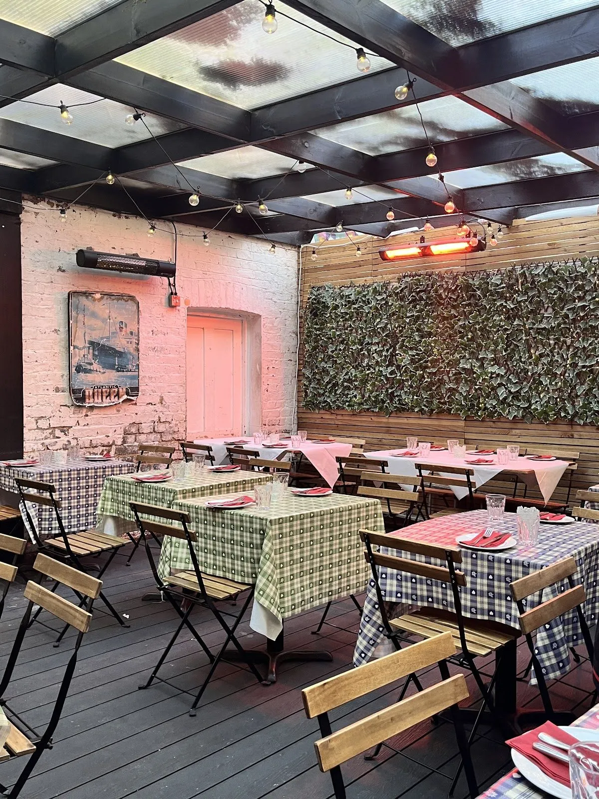 The covered garden room at Tsiakkos & Charcoal, with checked tablecloths, wooden folding chairs, a whitewashed brick wall, a hedge-covered wall and strings of fairy lights under a glazed roof