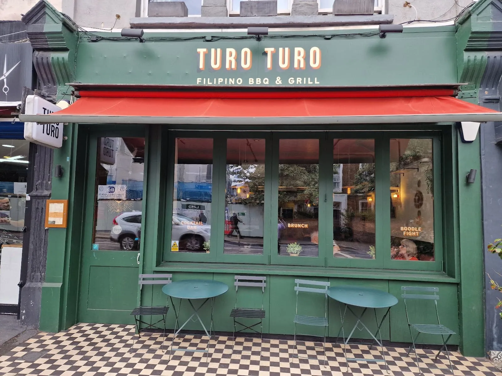 A green painted shopfront reading Turo Turo, Filipino BBQ and grill, under a red awning, with gold lettering across the windows and folding bistro tables on a black and white tiled pavement