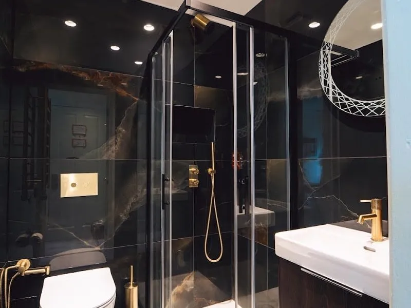 A black marble shower room at Twenty Nevern Square with a glass corner shower, brass taps and flush plate, and a white basin under a round mirror