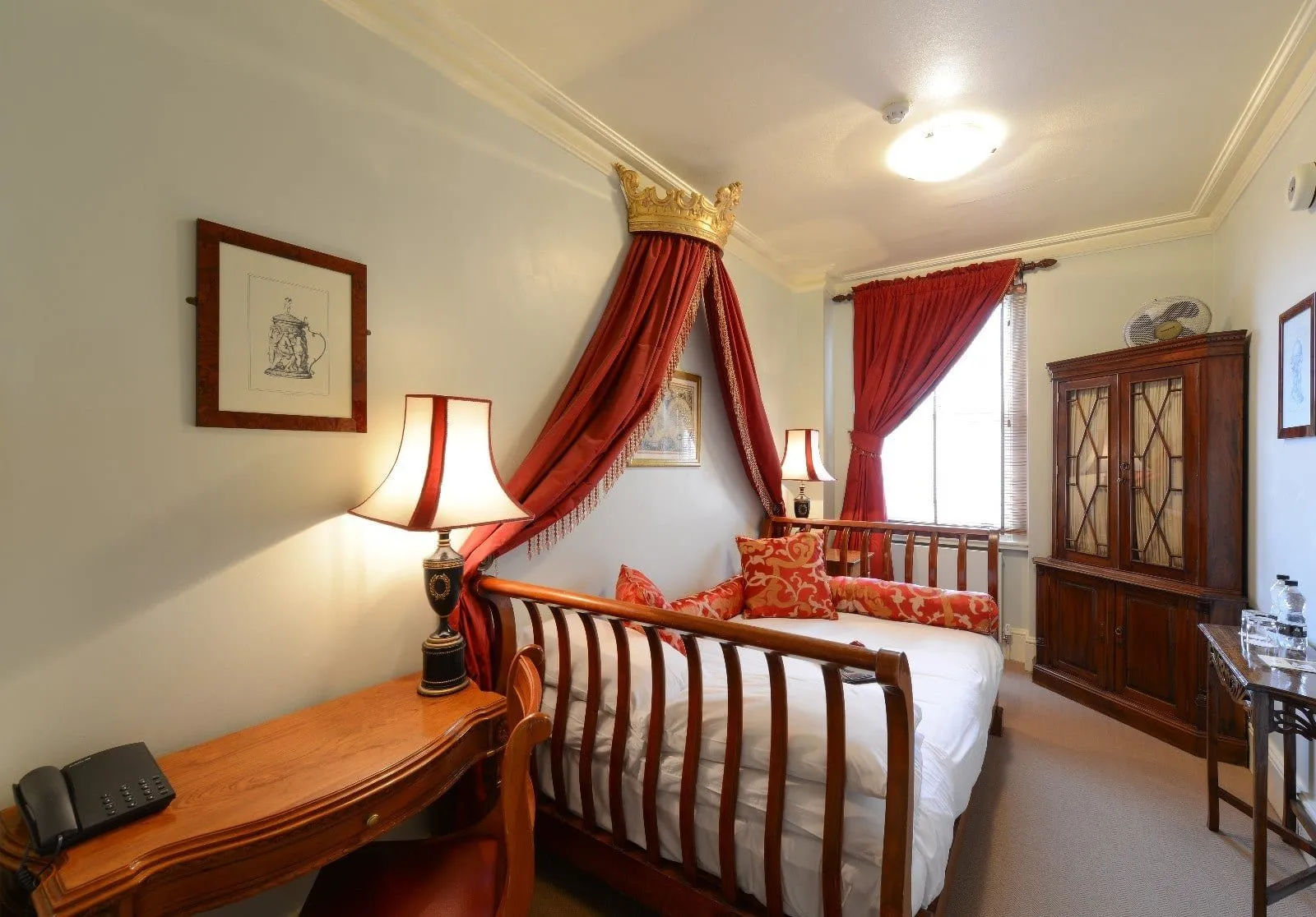 A Twenty Nevern Square bedroom with a wooden sleigh bed under a gilded crown and red silk drapes, red damask cushions, a carved desk and a glass-fronted mahogany cabinet by the window