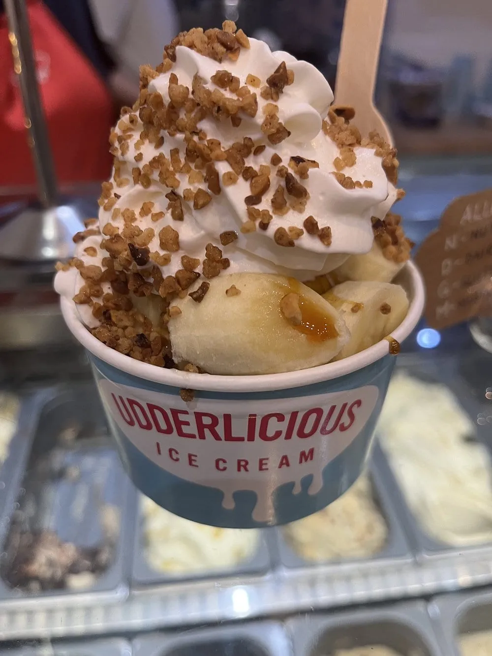 A tub of whipped ice cream loaded with nut brittle and banana slices, held over the shop's counter pans
