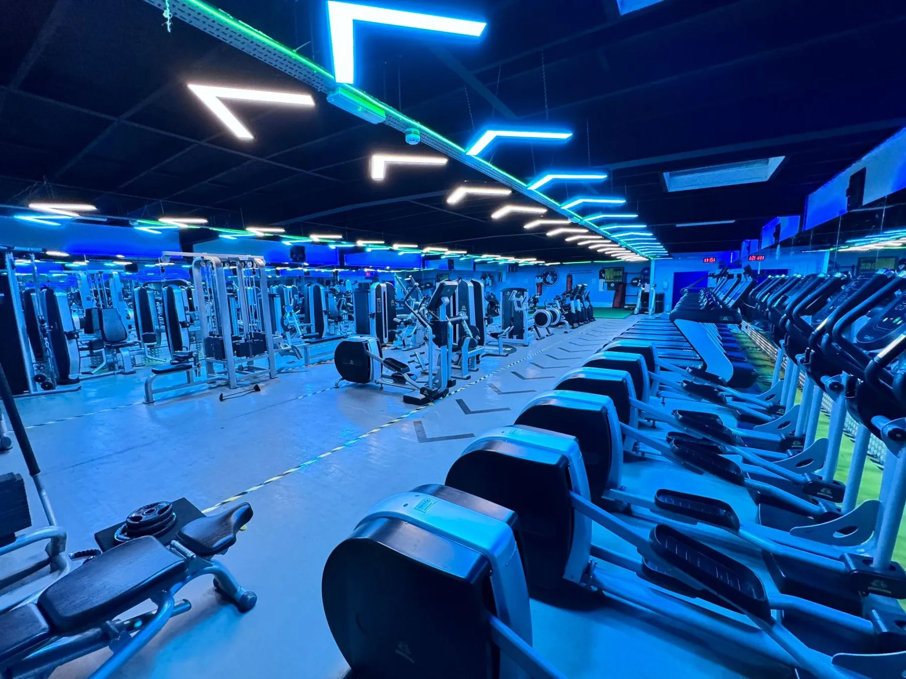 A long gym floor lit blue, neon chevrons across a black ceiling, cross-trainers in a row down one side and resistance machines down the other