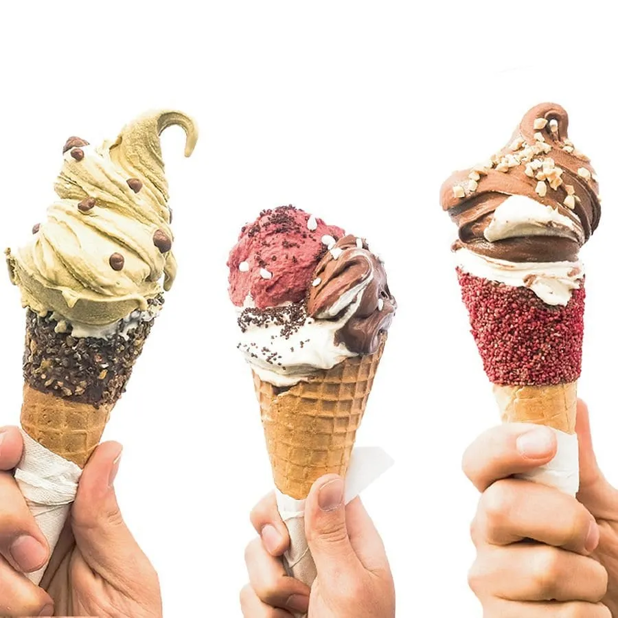 Three hands each holding an elaborately built cone — pistachio, berry with chocolate, and chocolate with chopped hazelnut — against a white background