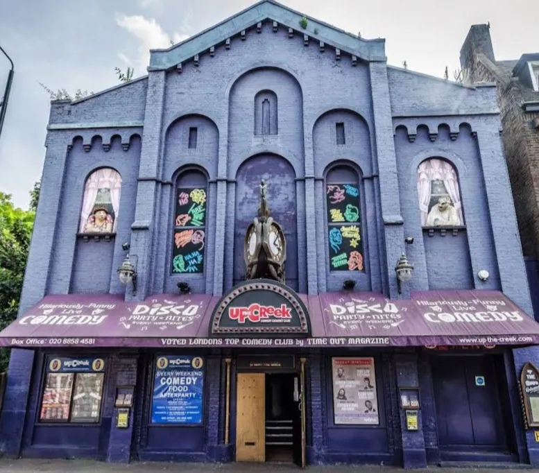 The purple, gothic-arched former church hall frontage of Up the Creek, with painted comedy banners and an awning reading "Disco Party Nites" and "Hilariously Funny Live Comedy"