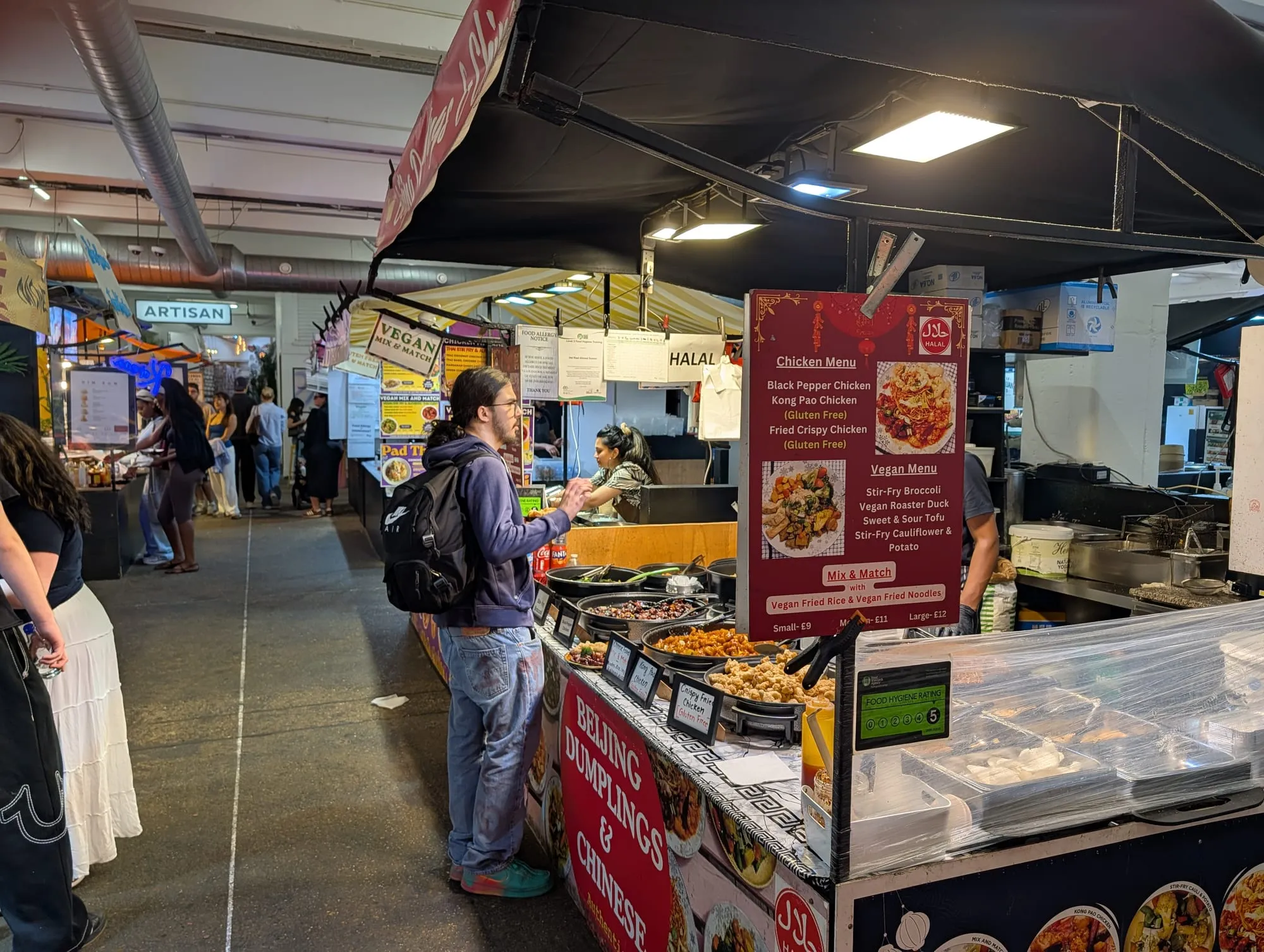 A stall at Upmarket on Brick Lane serving Beijing dumplings and Chinese food, its trays of crispy chicken and stir-fries under heat lamps, with hand-lettered signs and further stalls stretching down the hall behind