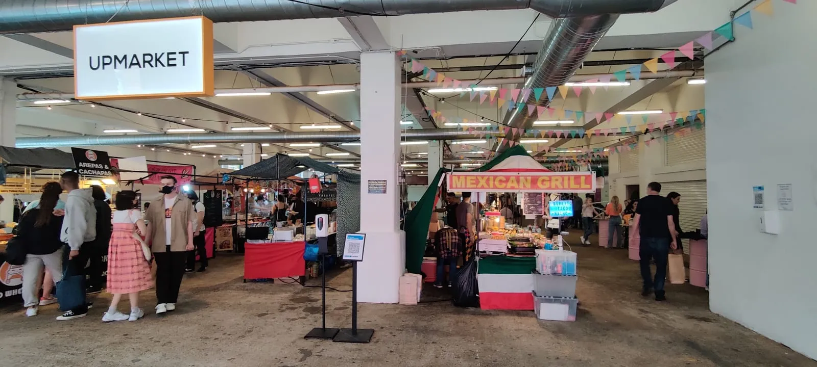 Street food stalls under strings of bunting inside Upmarket at the Old Truman Brewery, with the Upmarket sign hanging at the entrance