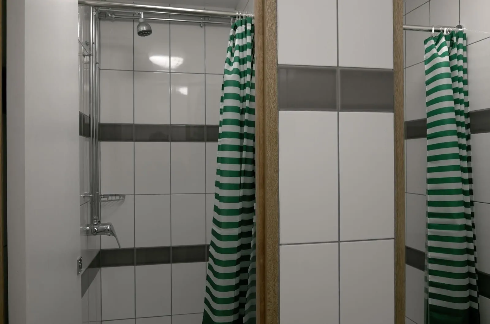Shared shower cubicles at Urbany Hostel London with white tiles, a grey band, wooden dividers and green and white striped curtains