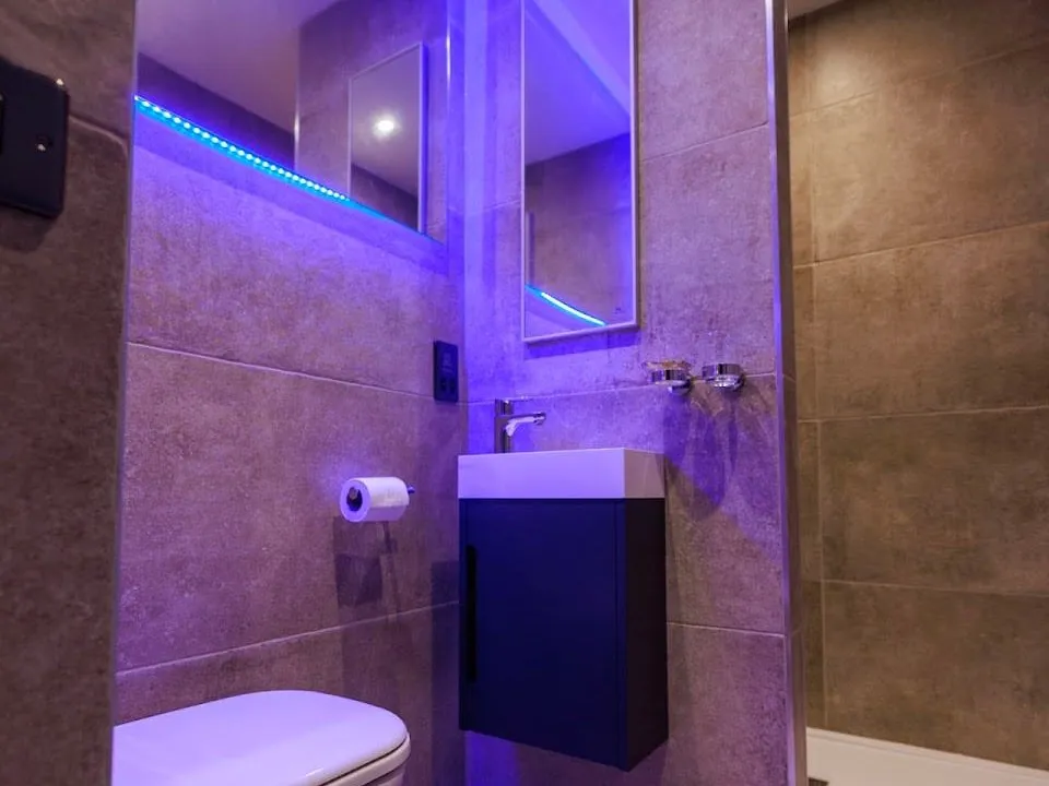 A small en-suite at Vancouver Hotel and Studios with grey stone-effect tiles, a wall-hung basin on a navy cabinet, a toilet and a mirror edged with purple LED light