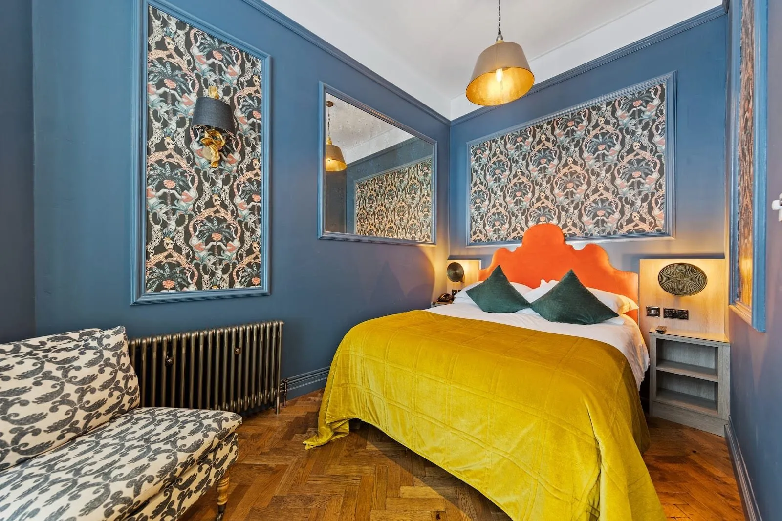 A room at Vancouver Hotel and Studios with blue walls, framed panels of monkey and palm-tree wallpaper, an orange velvet headboard, a mustard velvet bedspread, a patterned daybed and a parquet floor