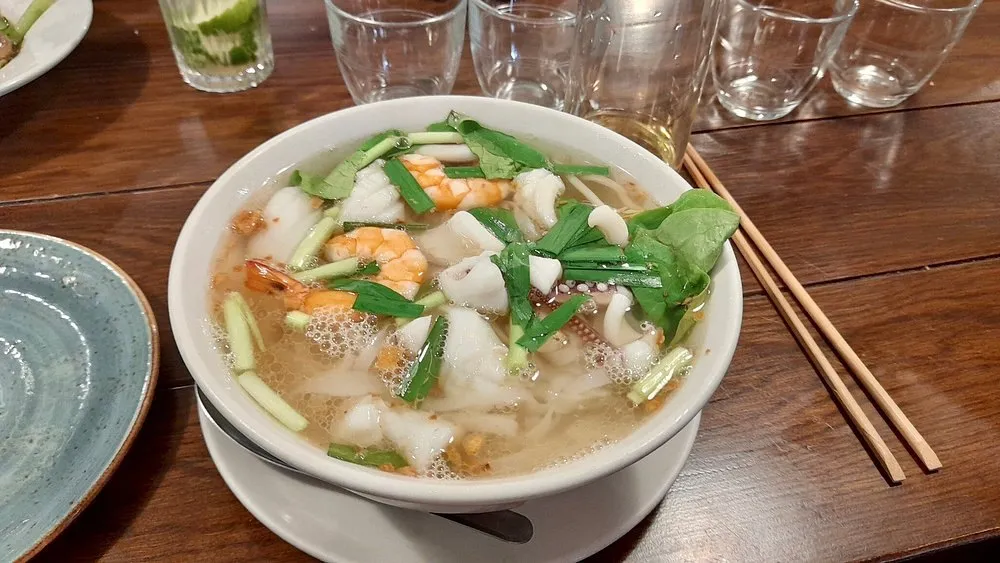 A bowl of clear Vietnamese noodle soup with prawns, squid, spring onion and herbs, on a wooden table with chopsticks alongside