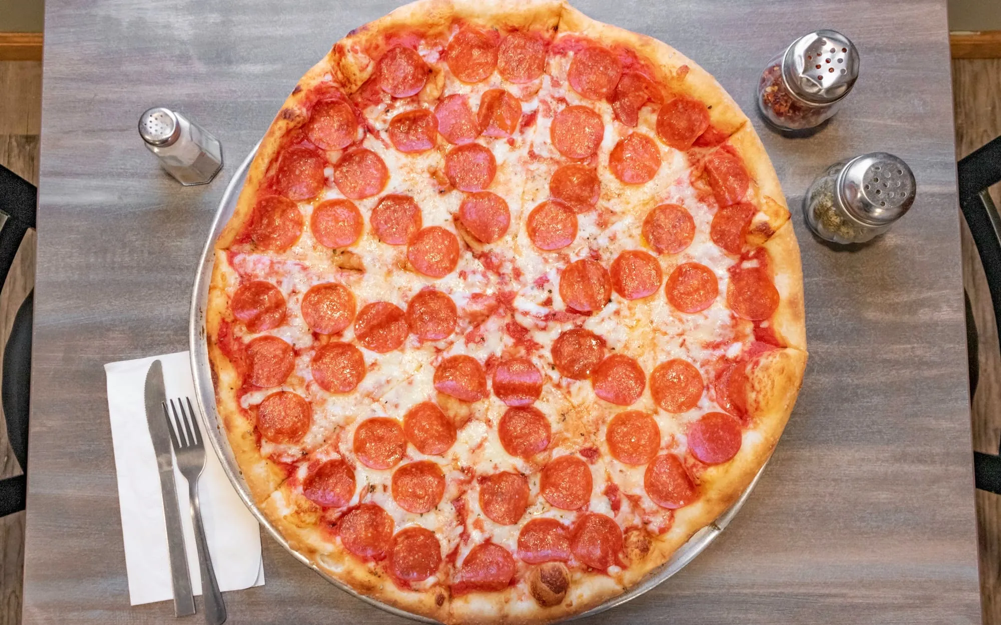A wide New York-style pepperoni pizza on a metal tray at Vincenzo's, cut into slices and covering most of the table, with shakers of chilli flakes and dried oregano beside it