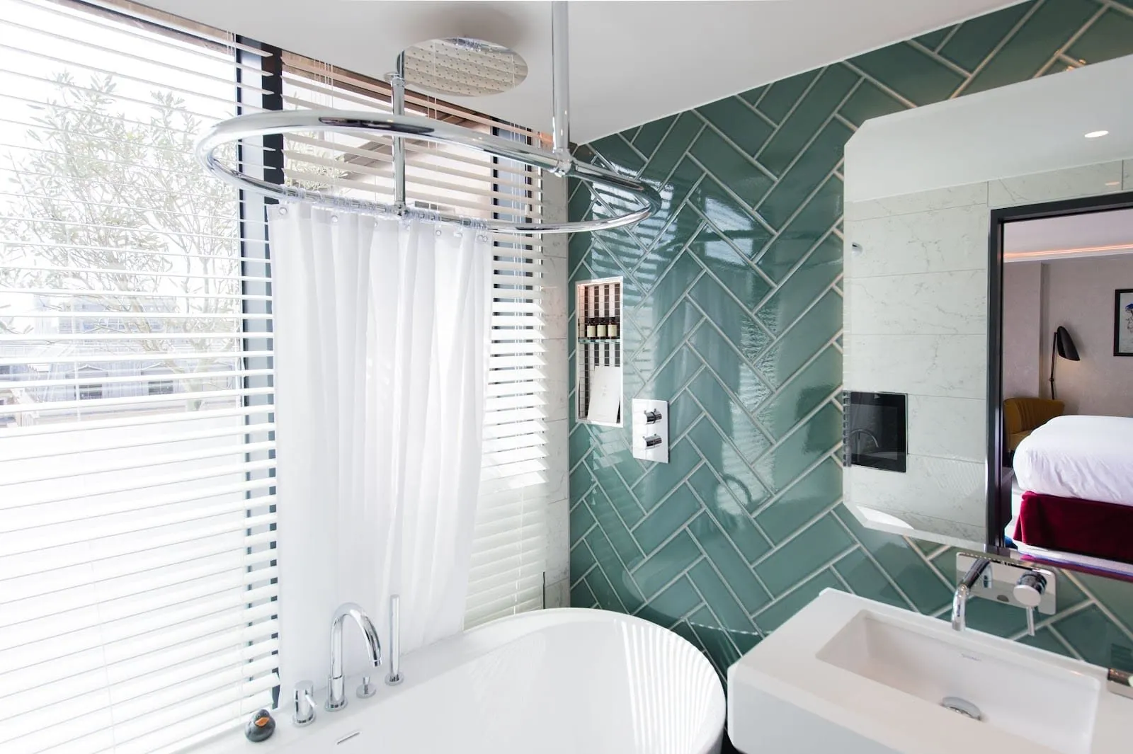 A bathroom at Vintry & Mercer with a freestanding white bath under a rain shower and curtain ring, a window with white blinds, and green herringbone tiles behind the basin
