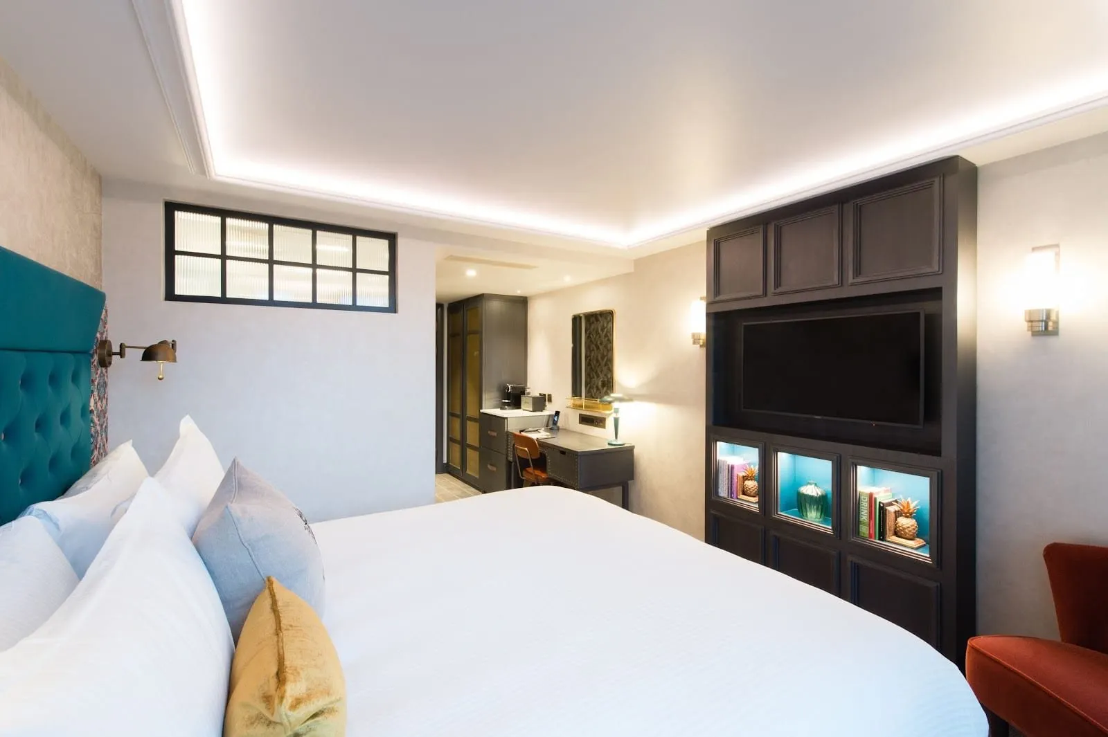 A bedroom at Vintry & Mercer with a teal buttoned-velvet headboard, a wide white bed, a dark wood cabinet holding the TV and lit teal shelves, a desk with a Nespresso machine and a rust-red armchair