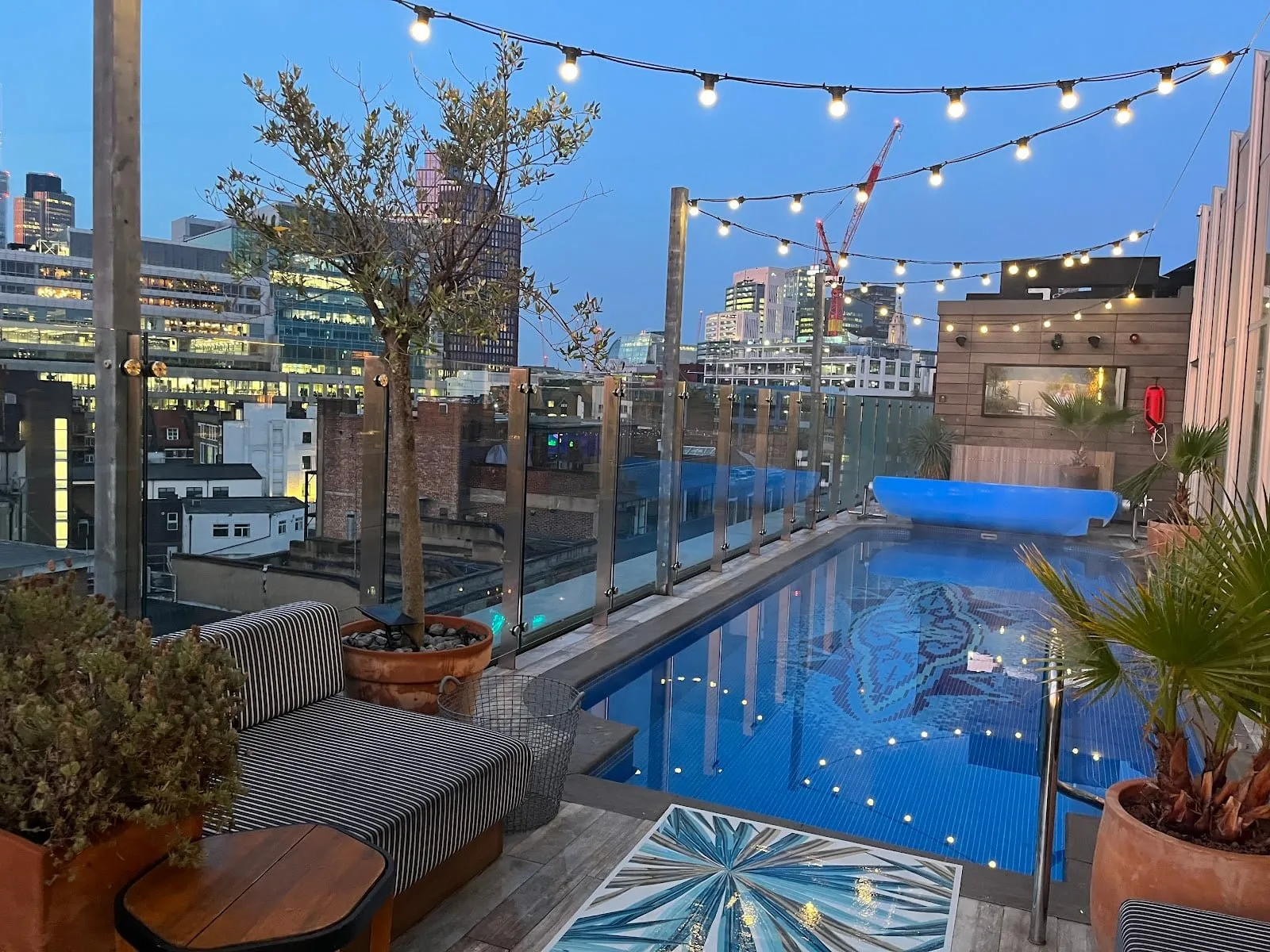 The rooftop pool at Virgin Hotels London-Shoreditch at dusk, under festoon lights, with the City towers beyond