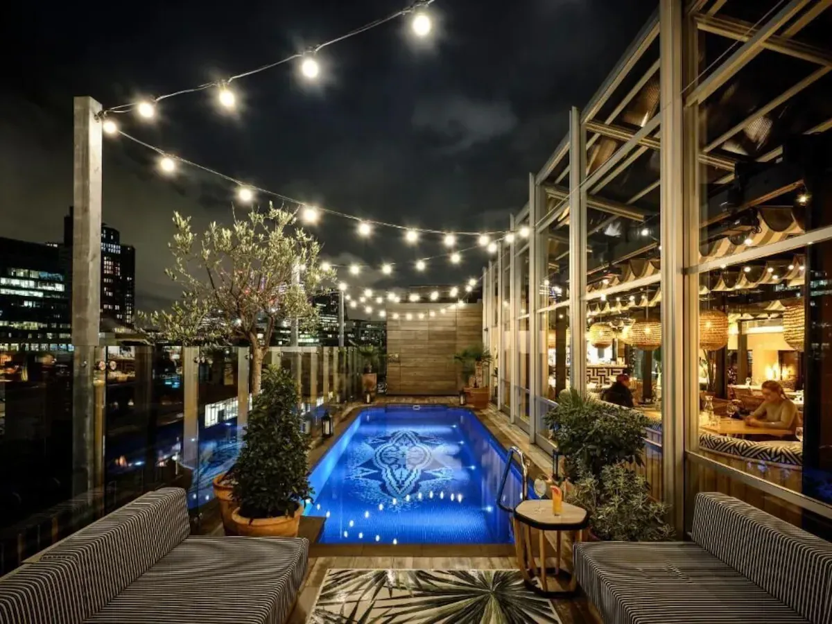 A lit rooftop pool at night with a mosaic patterned floor, festoon lights strung overhead, an olive tree in a pot and a glass-walled bar alongside, with City towers behind