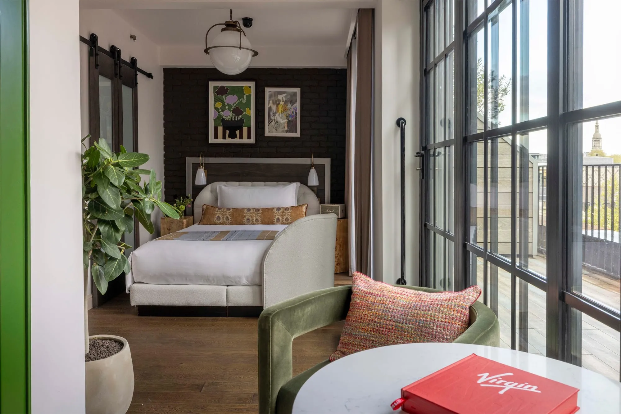 A room at Virgin Hotels London-Shoreditch with a dark brick accent wall, framed prints, an olive green armchair and floor-to-ceiling windows opening onto a terrace