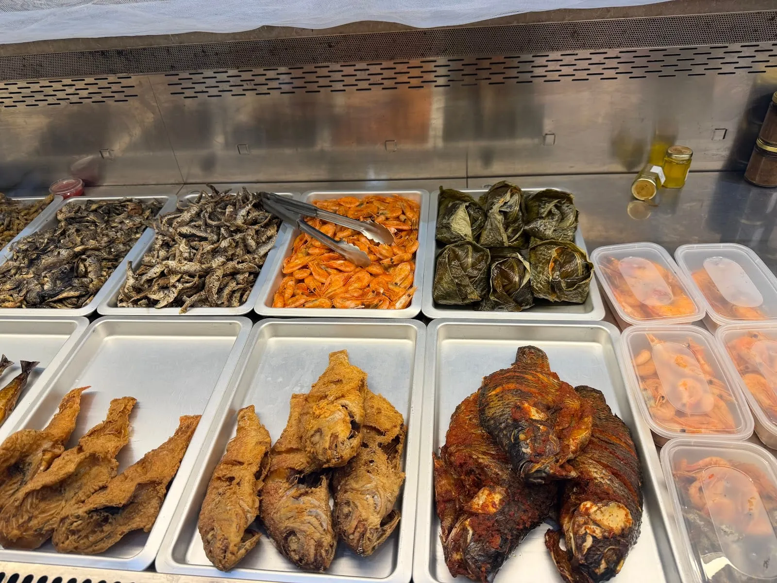 A Ghanaian hot counter in stainless trays: two trays of small dried fish, a tray of cooked orange prawns with tongs in them, a tray of leaf-wrapped parcels, golden deep-fried fish, and whole fish in a dark red stew, with plastic tubs of prawns in sauce stacked to one side