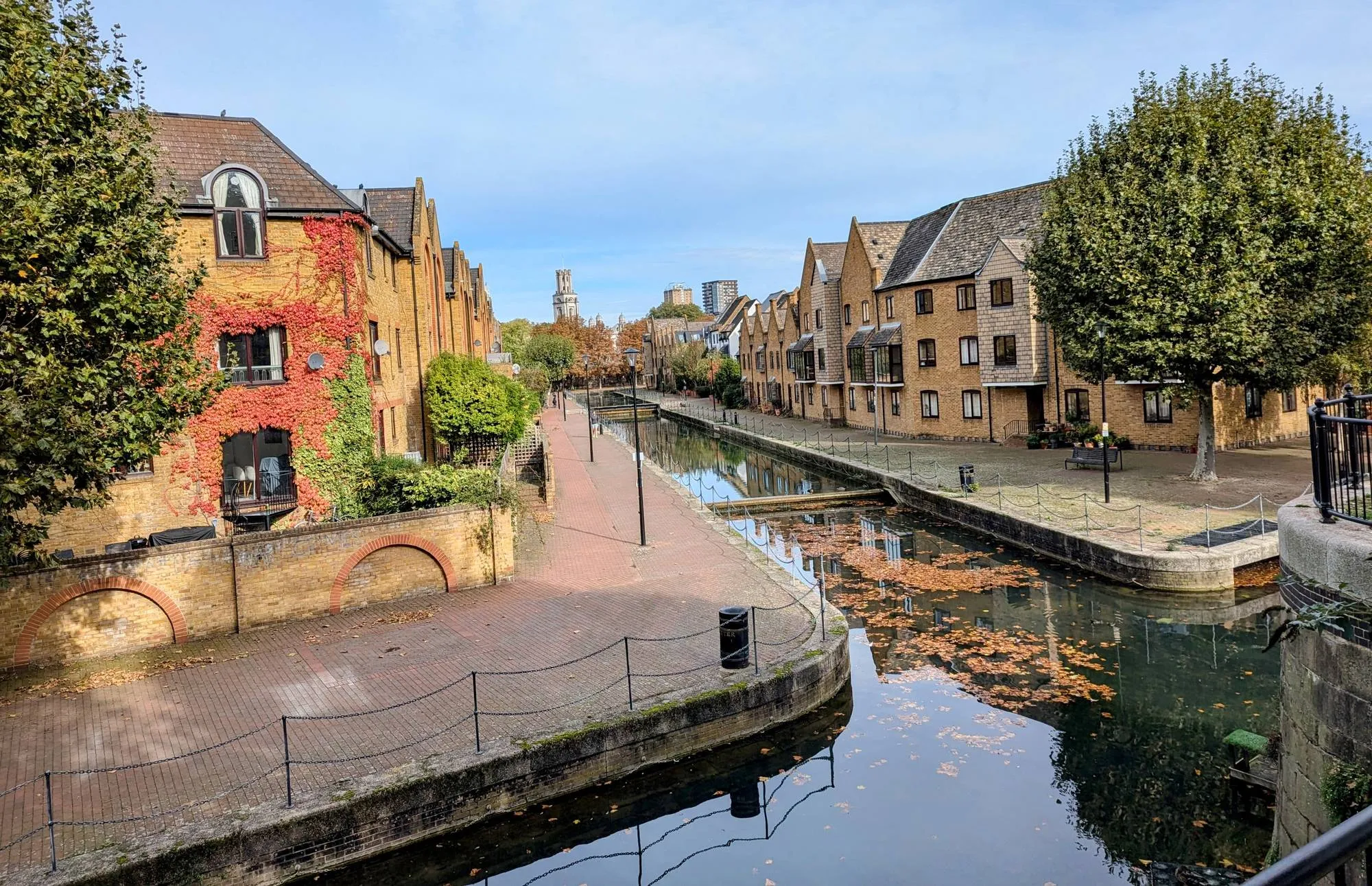 The Ornamental Canal in Wapping, with still water reflecting autumn leaves, brick warehouse-style housing on both banks and a church tower in the distance