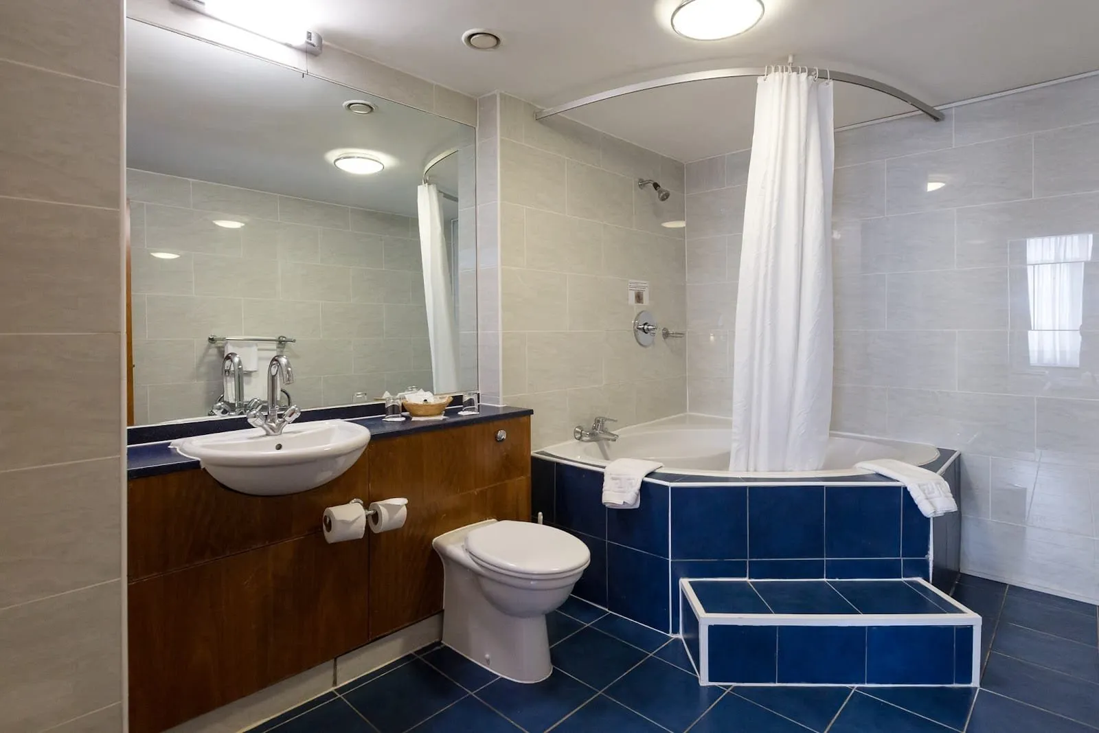 A bathroom at the Wembley International Hotel with a corner bath boxed in navy tiles, a step up to it, a curved shower curtain rail and a basin set in a wood-panelled unit