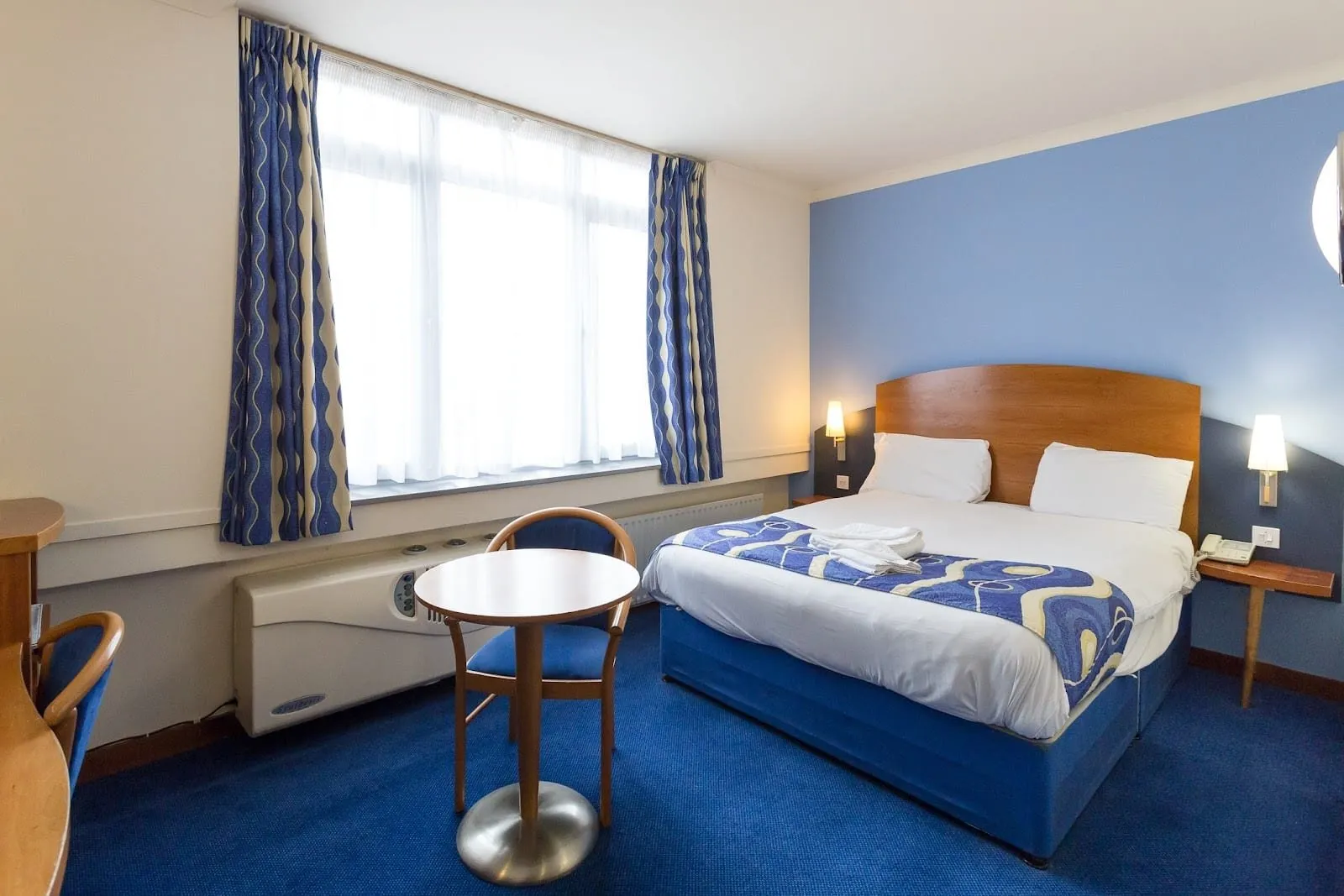 A double room at the Wembley International Hotel with a blue carpet, a light blue wall behind a wooden headboard, blue patterned curtains and an air-conditioning unit under a wide window