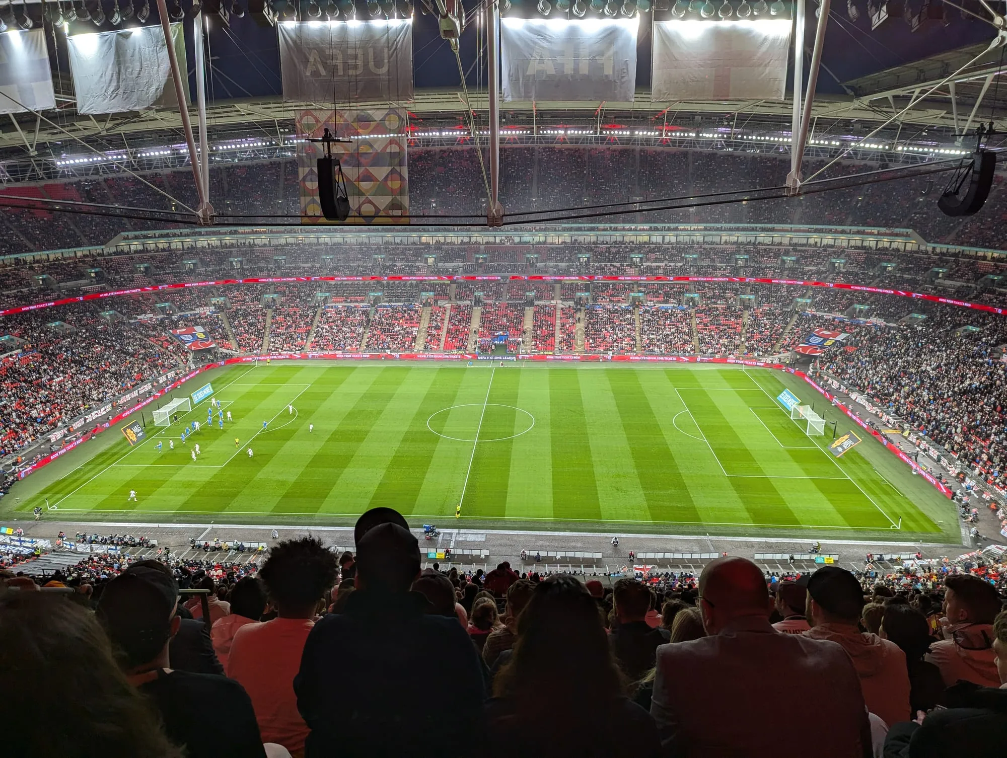 The inside of Wembley Stadium during an evening England match, the pitch floodlit and the tiers full, seen from high in the upper bowl