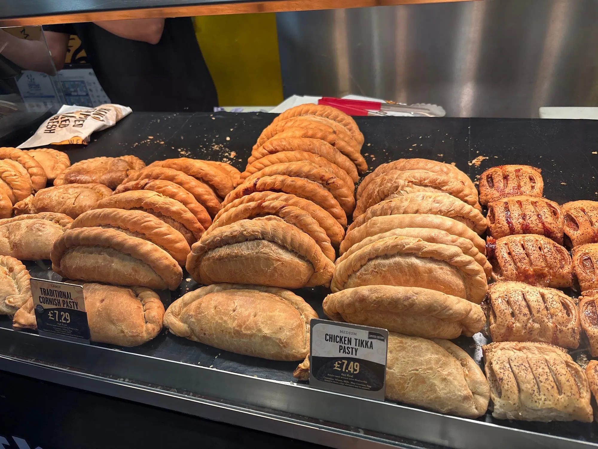 Golden Cornish pasties with crimped edges in a hot-food display case, labelled Traditional Cornish Pasty.