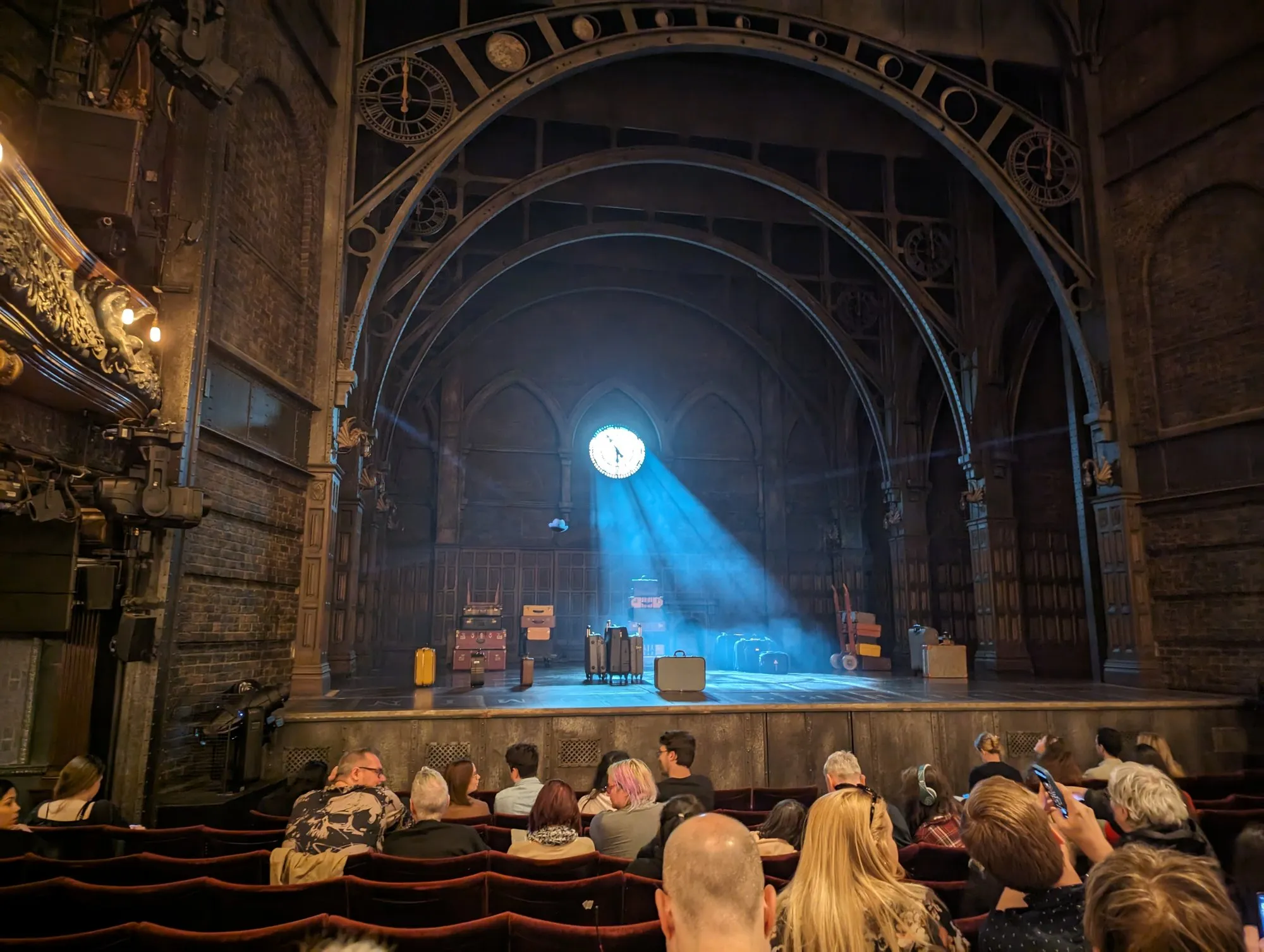 The stage of the Palace Theatre set for Harry Potter and the Cursed Child, gothic arches and clock faces above a pile of trunks and suitcases lit by a blue shaft of light