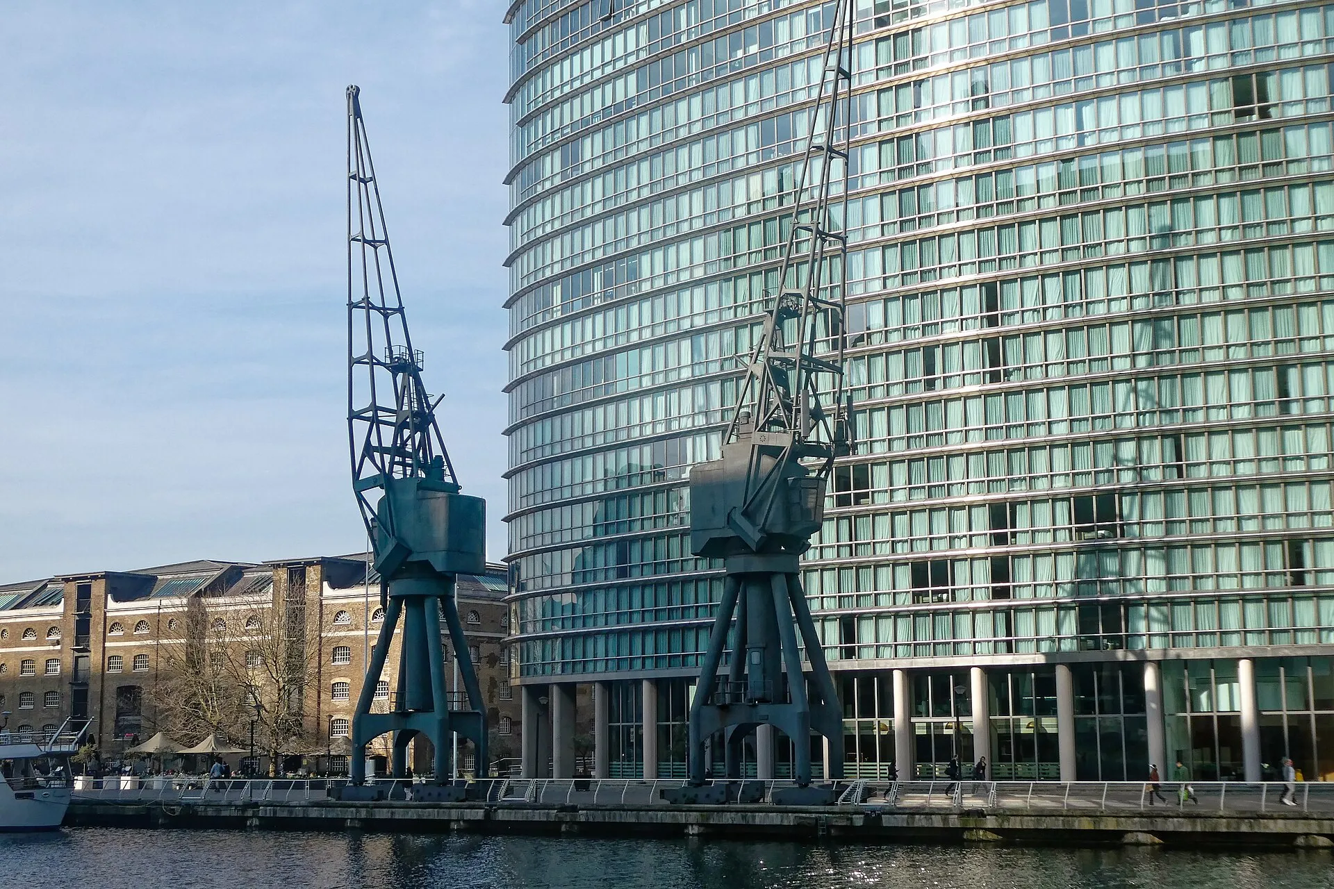 Two preserved dockside cranes on West India Quay in front of the curved glass tower of the London Marriott Hotel Canary Wharf, with the brick Georgian warehouses of the quay on the left and the dock in the foreground