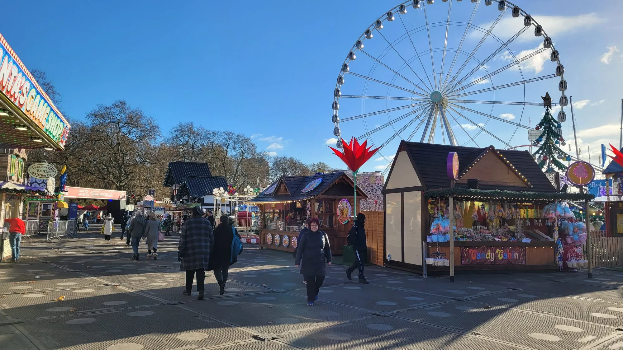 The observation wheel at Winter Wonderland under a clear blue sky, with a candy stall, a games stall and a decorated Christmas tree along an almost empty walkway