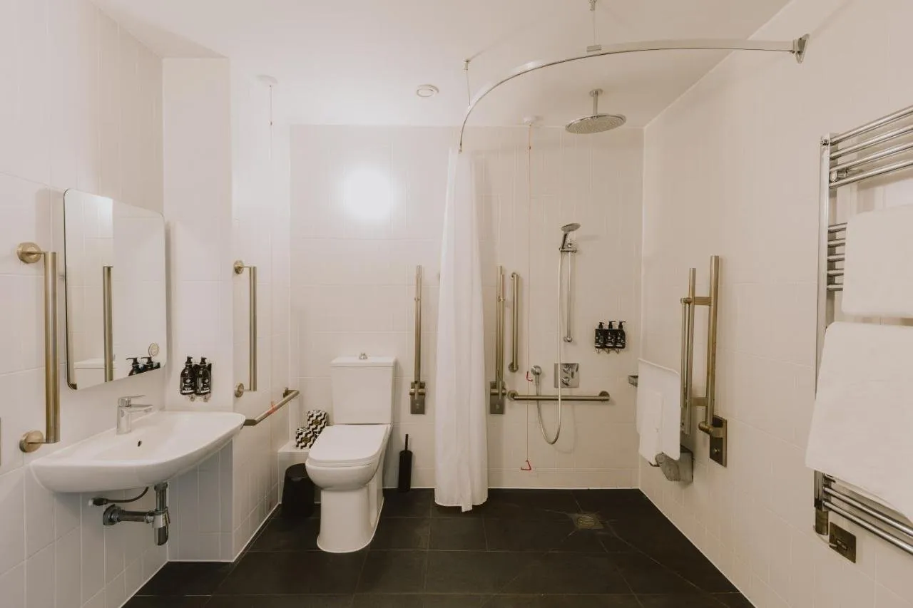 An accessible wet-room bathroom at Wilde Aparthotels London Paddington with a level-floor shower behind a curtain, brass grab rails, a fold-down shower seat and dark floor tiles