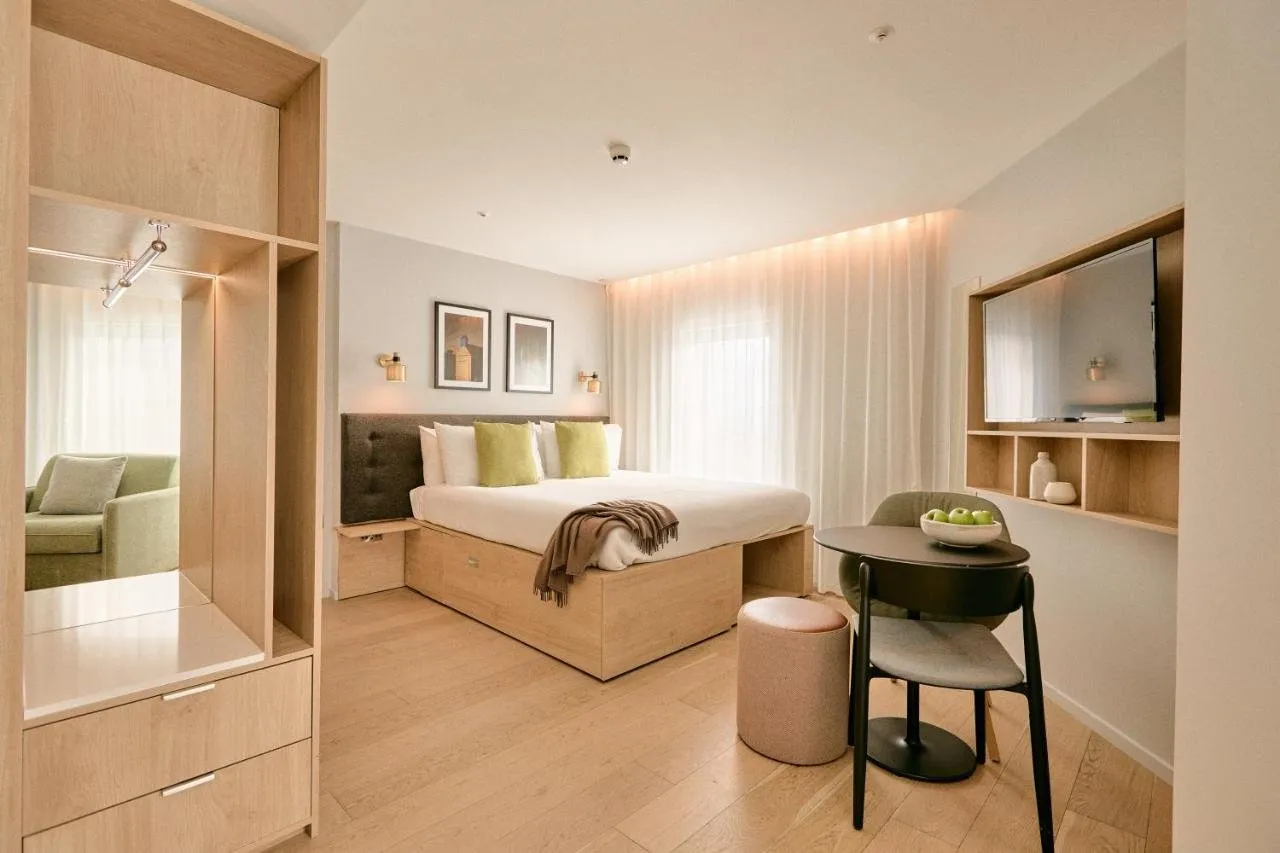 A studio at Wilde Aparthotels London Paddington with a double bed on an oak storage base, an open oak wardrobe with a mirror, a small round table with two chairs and floor-length voile curtains