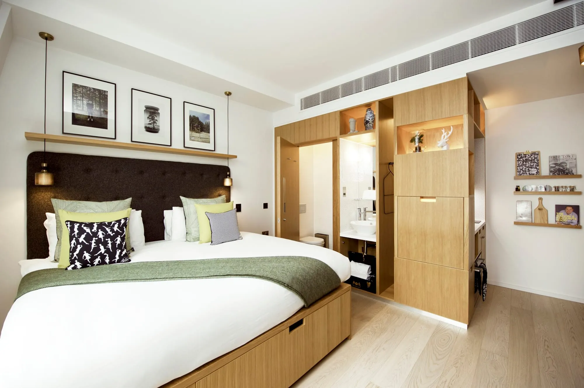 A studio at Wilde Covent Garden, with a dark tufted headboard, framed photography above the bed and an open wood-panelled unit holding storage and the ensuite