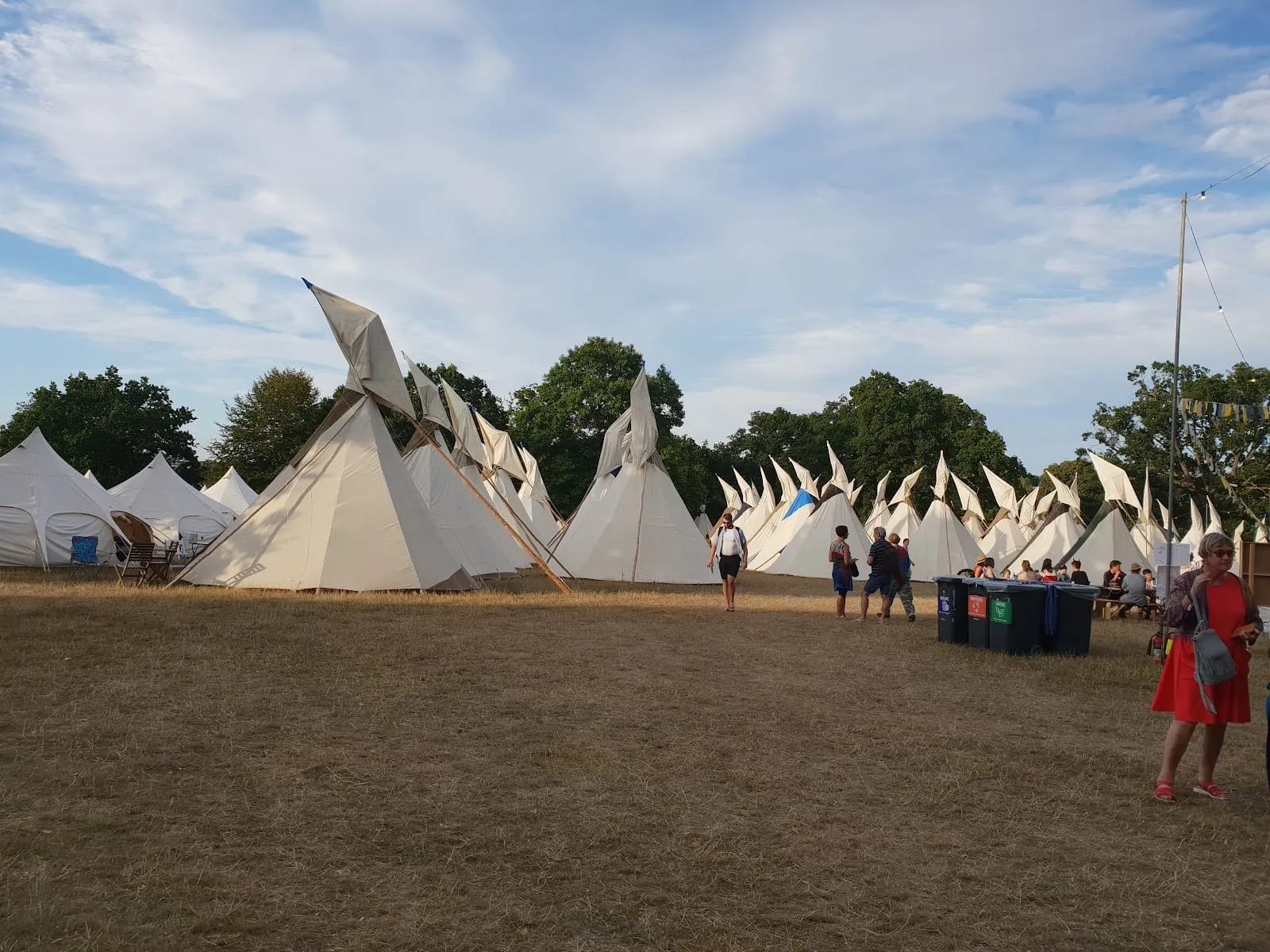 White canvas tipis and bell tents in a sunlit field of dry grass, with people walking between them