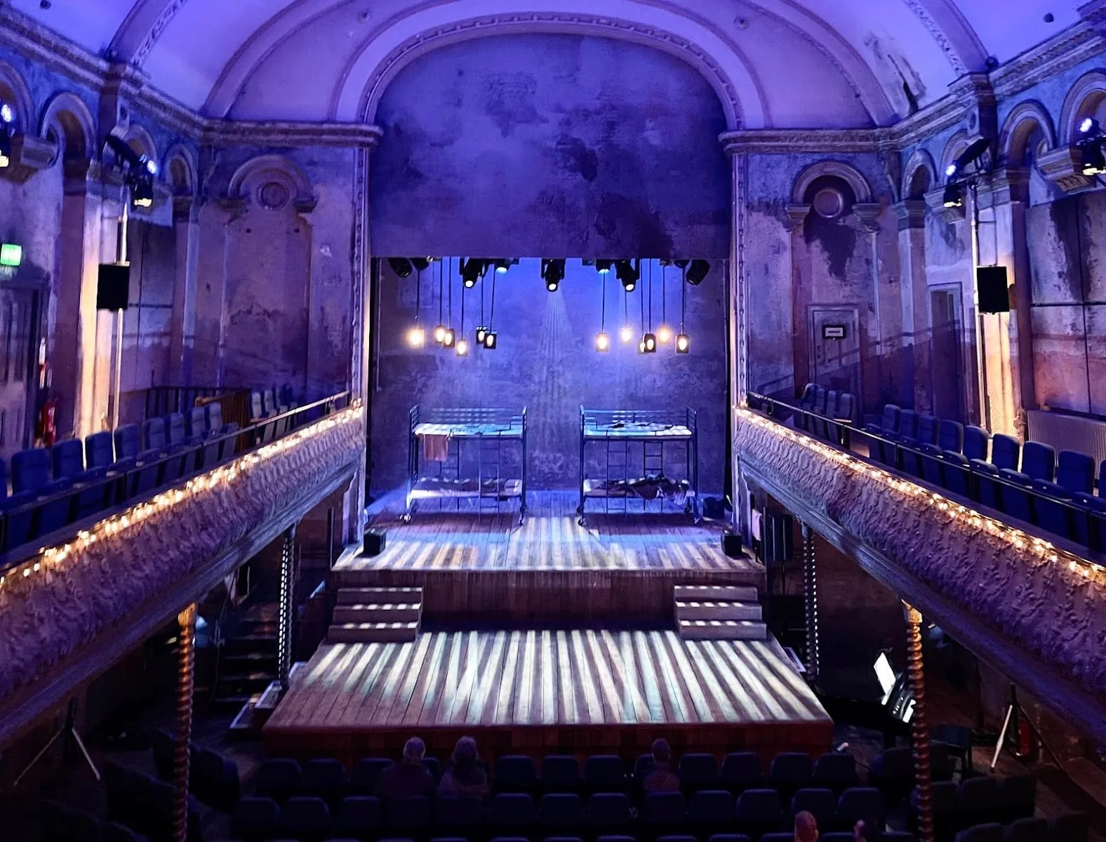 Wilton's Music Hall in blue light, with fairy lights along the balcony fronts, twisted columns beneath and a wooden stage under a high arched ceiling