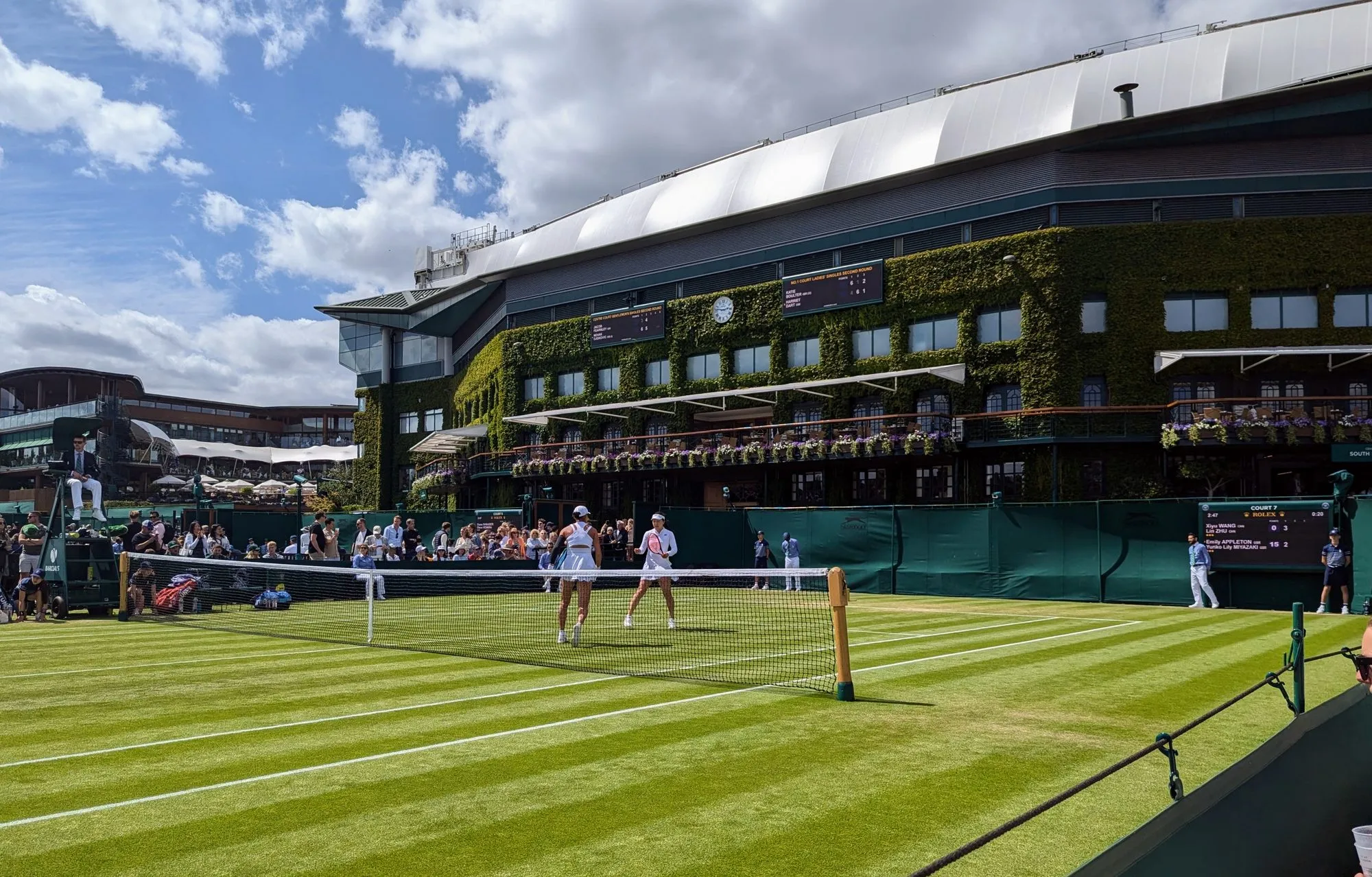 A ladies' doubles match on an outside court at Wimbledon, with the ivy-clad Centre Court building behind