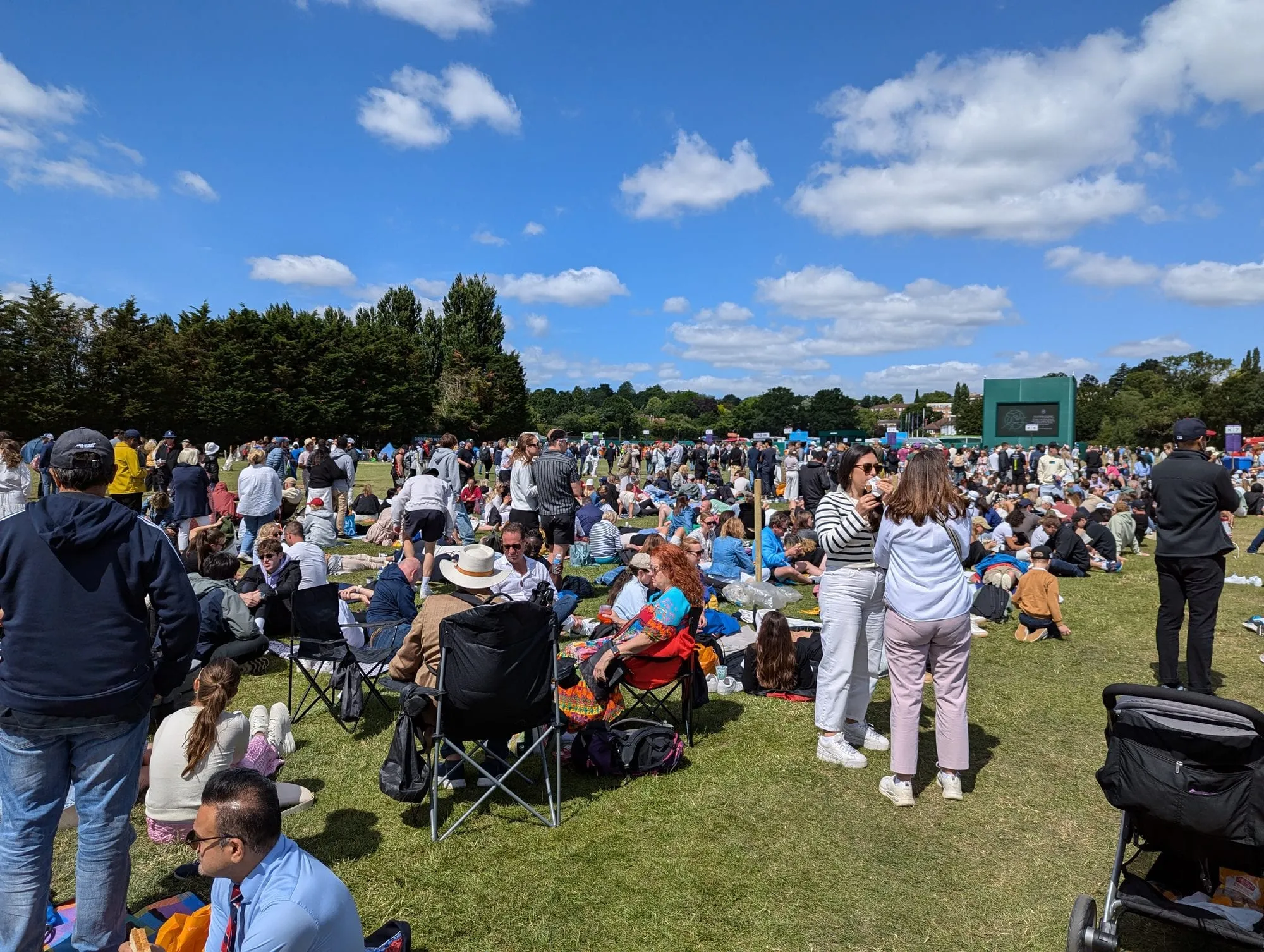 A large crowd waiting on the grass in Wimbledon Park during The Championships, with camping chairs, picnic blankets, queue section marker boards and a big screen in the distance