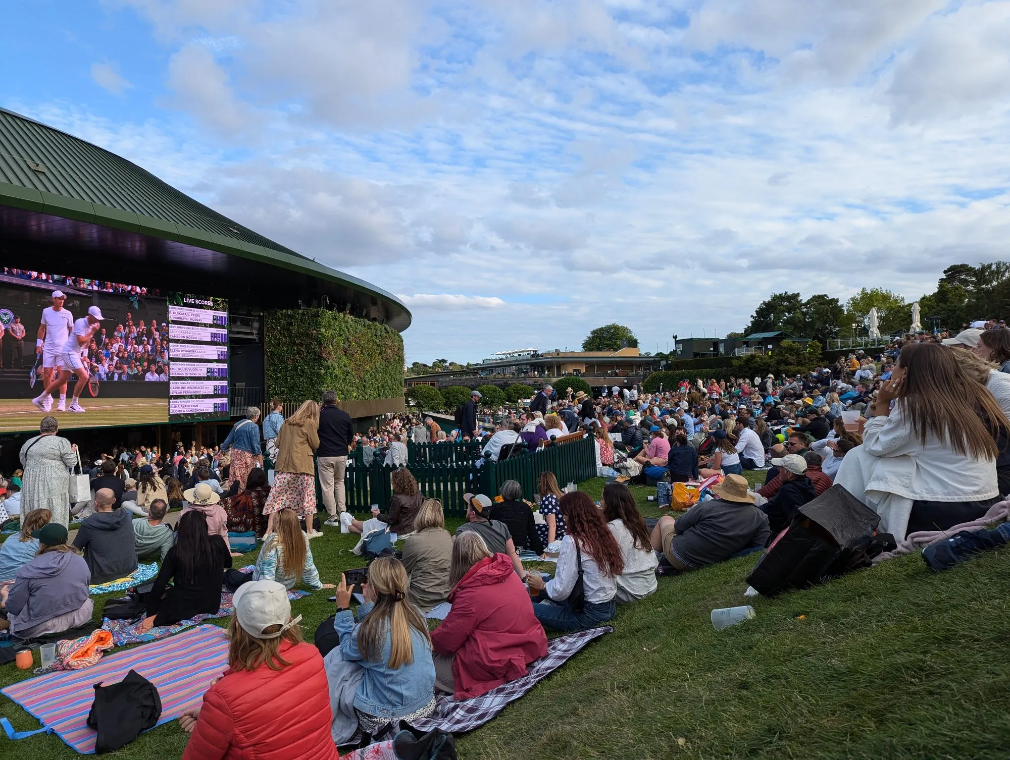 Spectators sitting on the grass of The Hill at Wimbledon watching the big screen on the side of No.1 Court in the evening