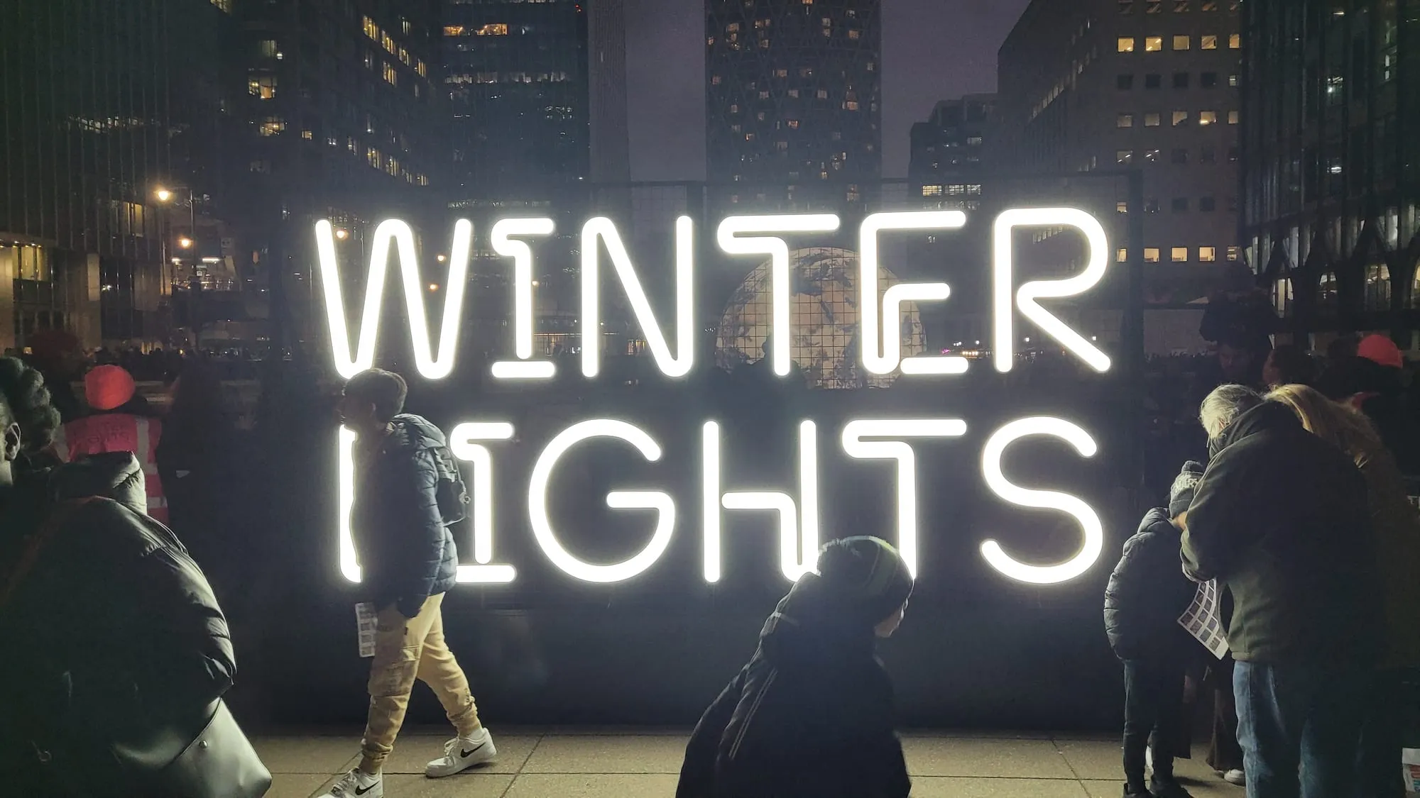 A large illuminated WINTER LIGHTS sign at Canary Wharf at dusk, with people walking past