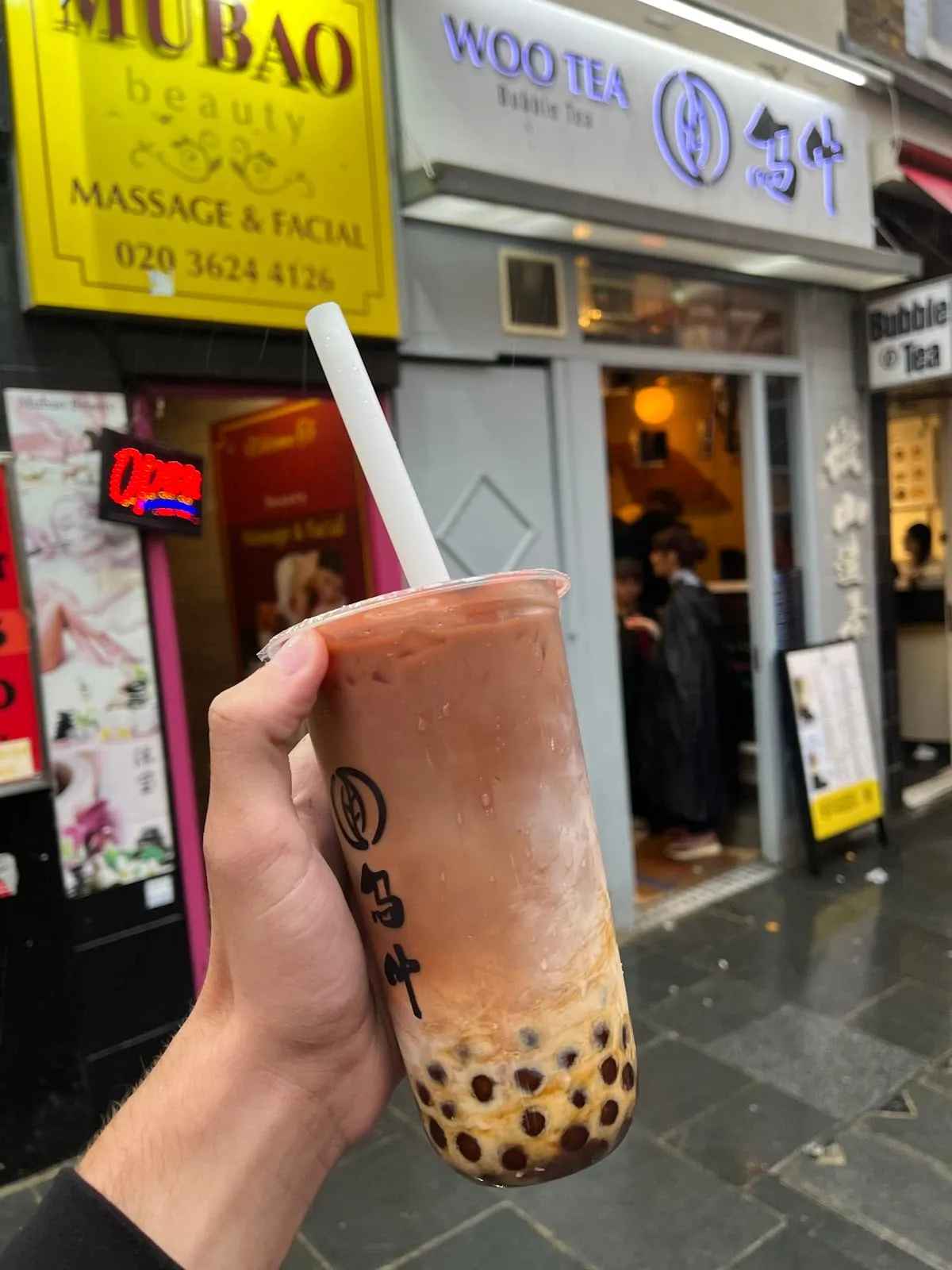 A Woo Tea milk tea with brown sugar pearls held up on the pavement outside the shop's sign
