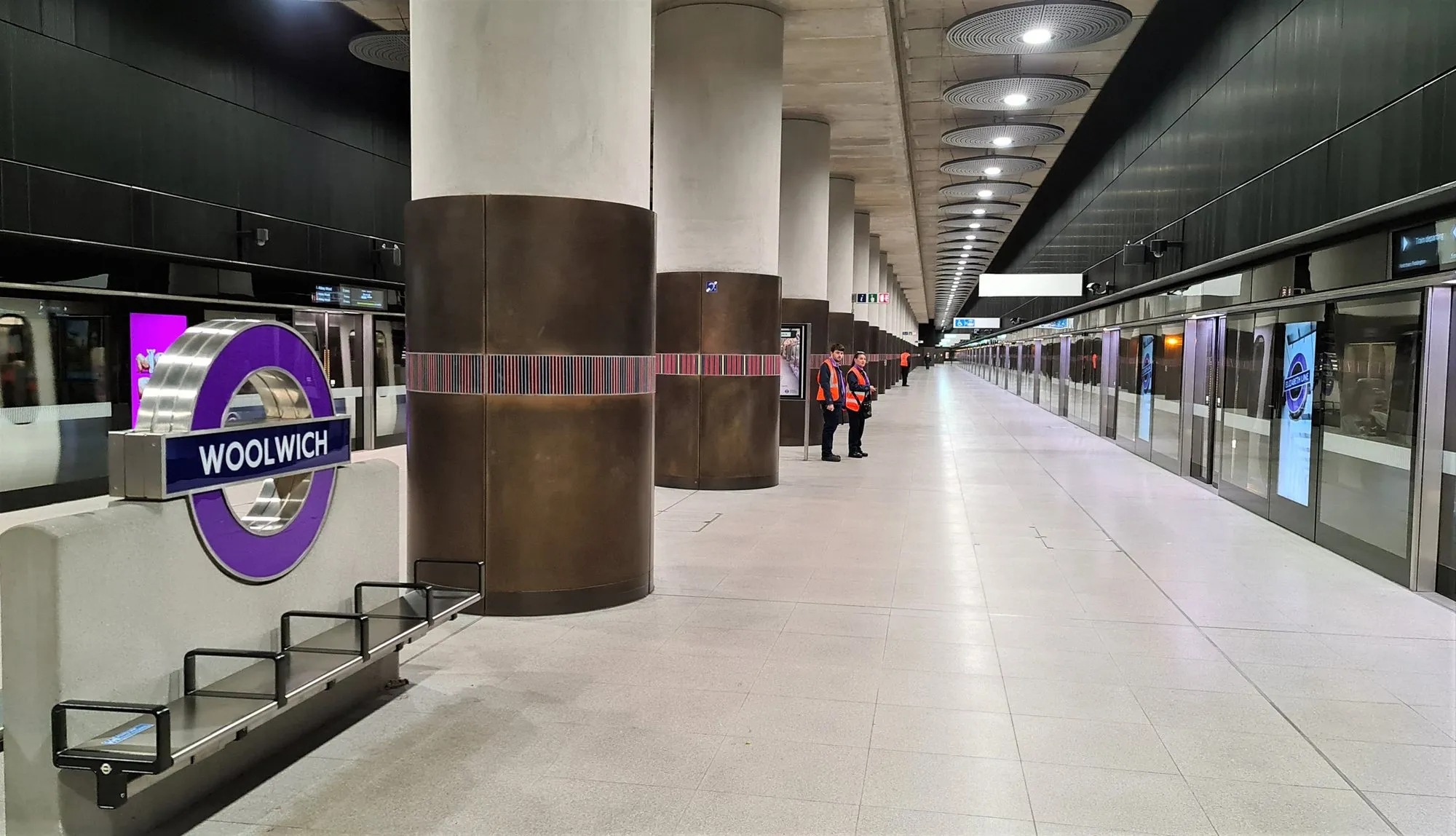The Elizabeth line platform at Woolwich: a wide flat concourse with platform-edge doors along the right-hand side, a train at the far platform and two staff in high-visibility jackets