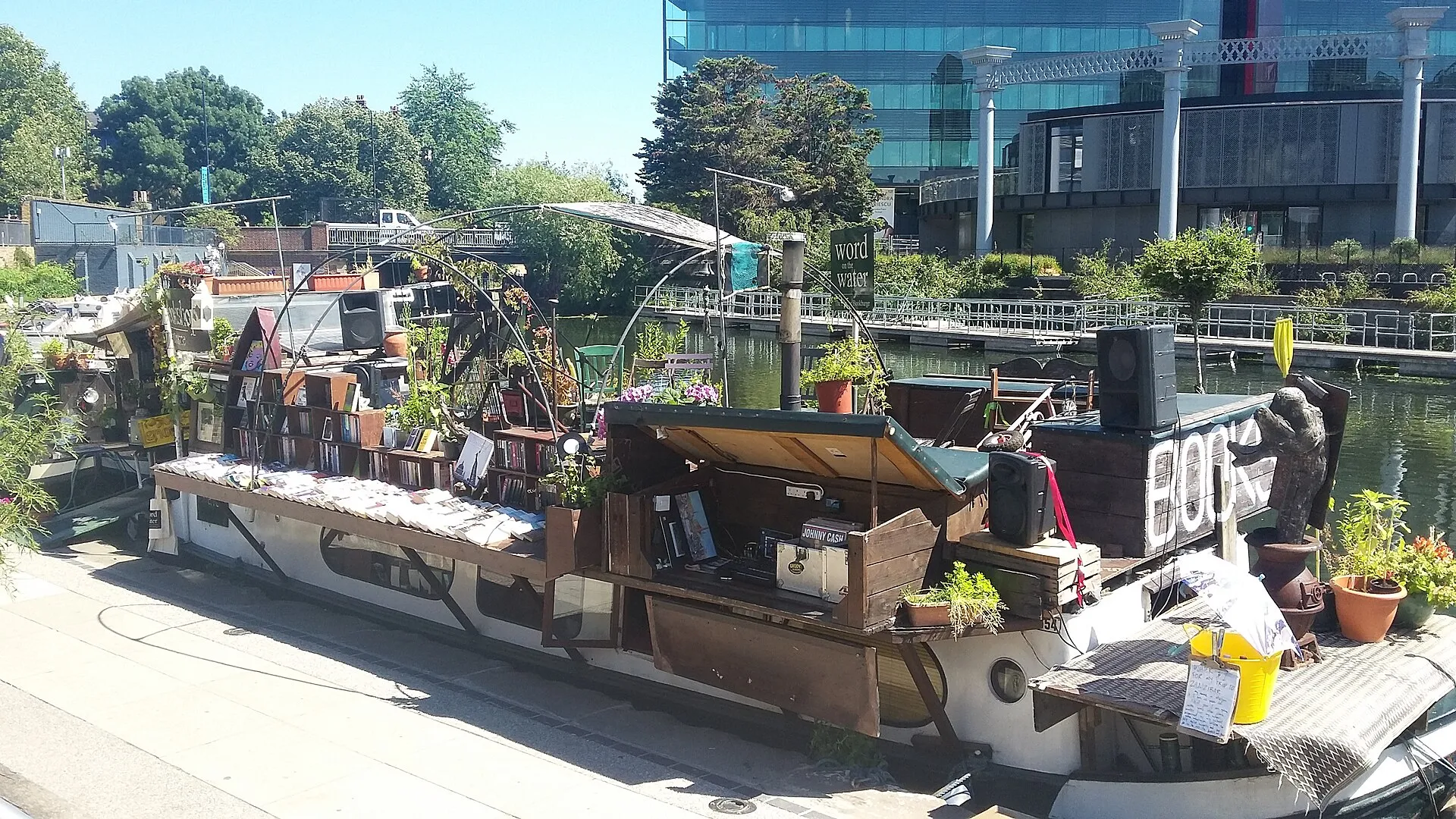 The Word on the Water bookshop barge moored on the canal, with books laid out along the roof and a deck of plants and speakers