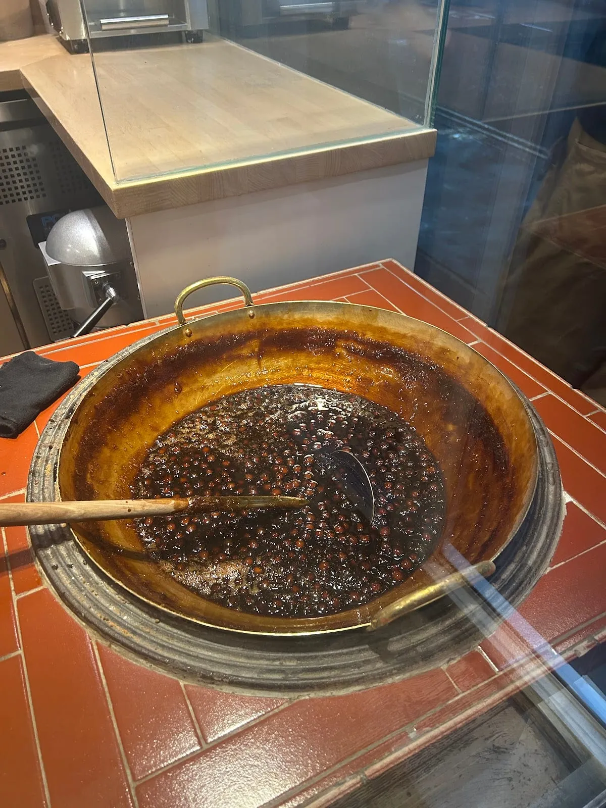 A wide wok of brown sugar tapioca pearls simmering in dark syrup behind a glass screen, with a wooden paddle resting in it