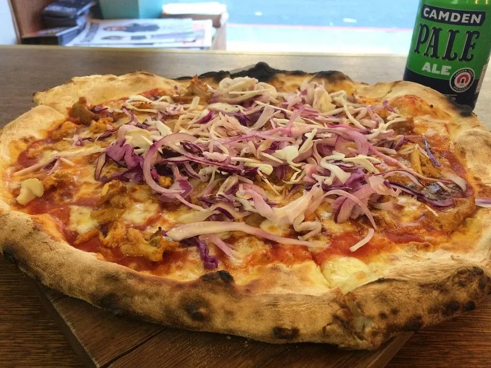 A Yard Sale pizza on a wooden table piled with shredded red and white cabbage slaw, beside a can of Camden Pale Ale
