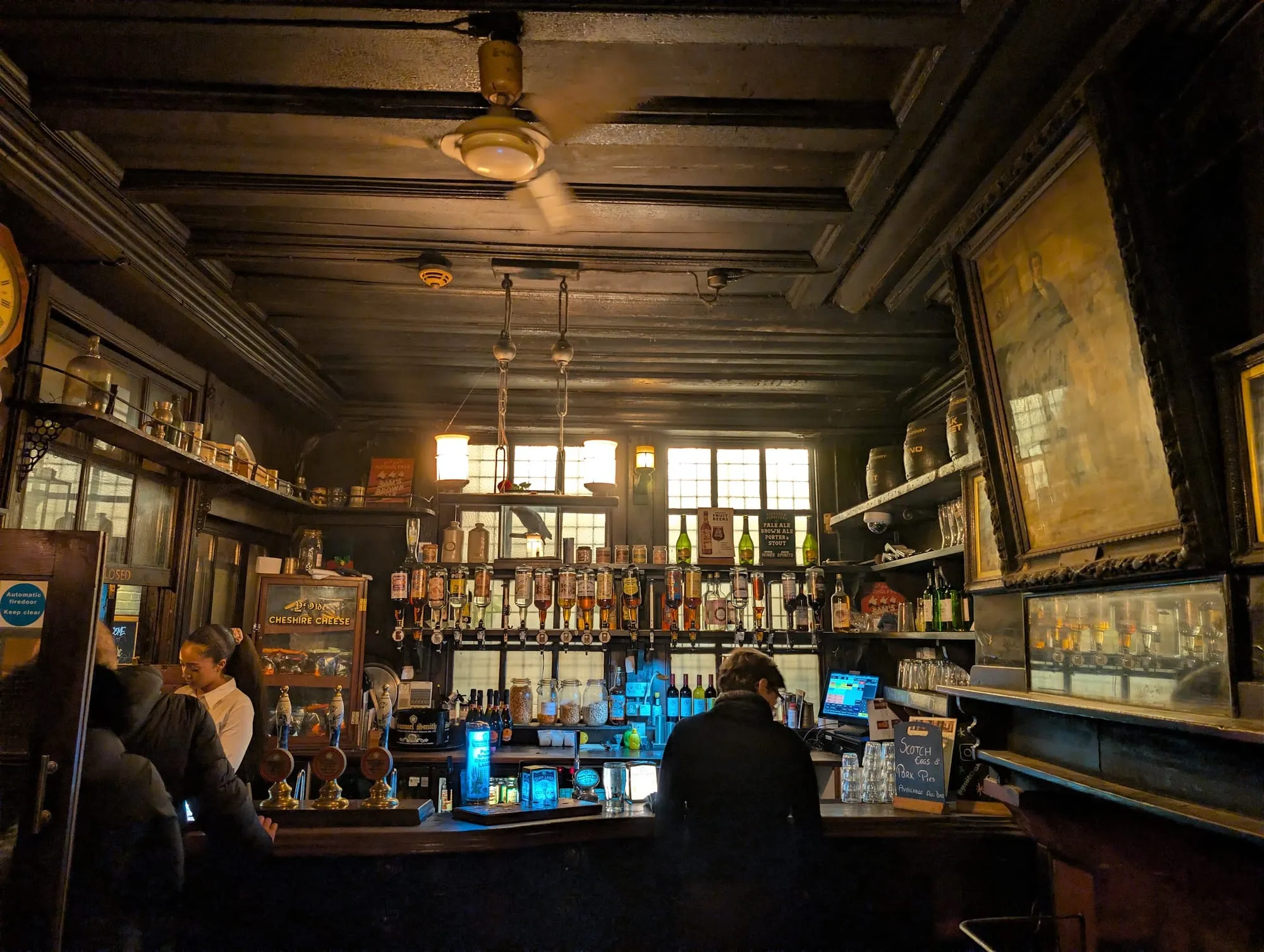 A dim, low-beamed bar at Ye Olde Cheshire Cheese, with dark wood fittings, hanging lamps and framed portraits on the walls