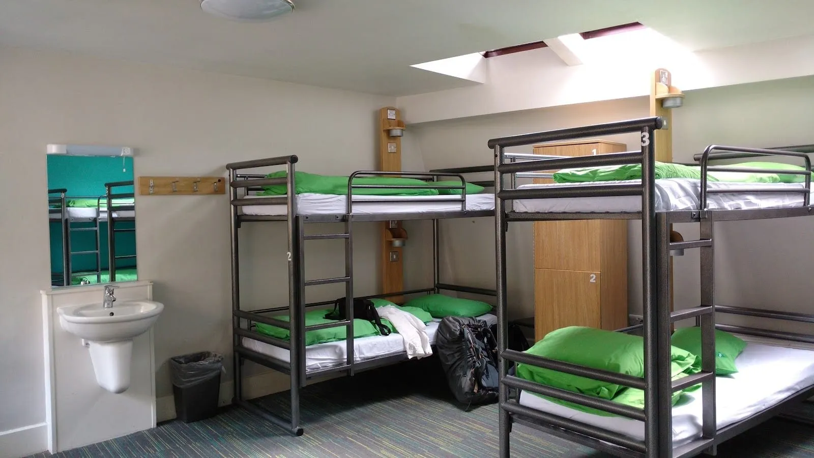 A dormitory at YHA London St Paul's with numbered grey steel bunk beds made up in green bedding, tall wooden lockers, a skylight and a washbasin with a mirror on the wall