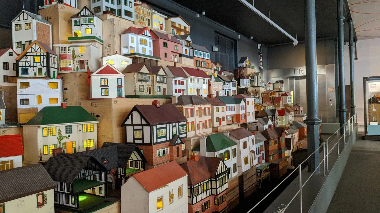 A wall-mounted display of dozens of miniature dolls' houses in different architectural styles, lit from within, at Young V&A