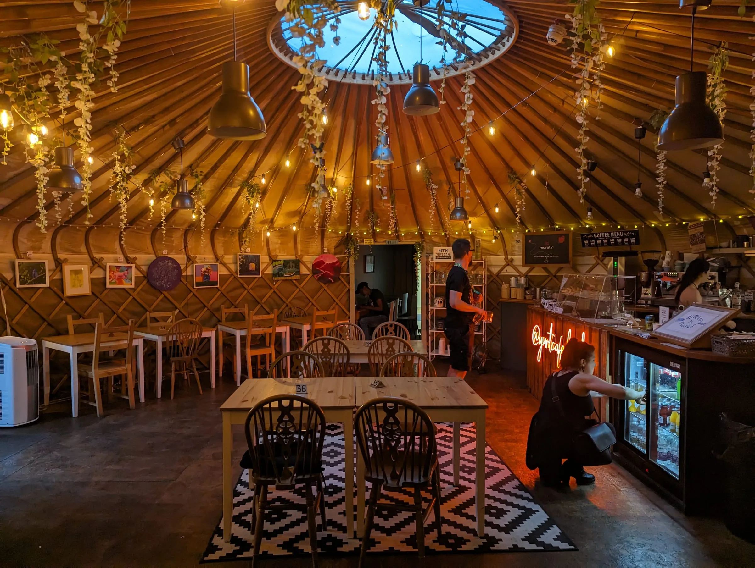 Inside the Yurt Café: a round timber-ribbed ceiling hung with lights and flowers, wooden tables on a patterned rug and the counter at the back